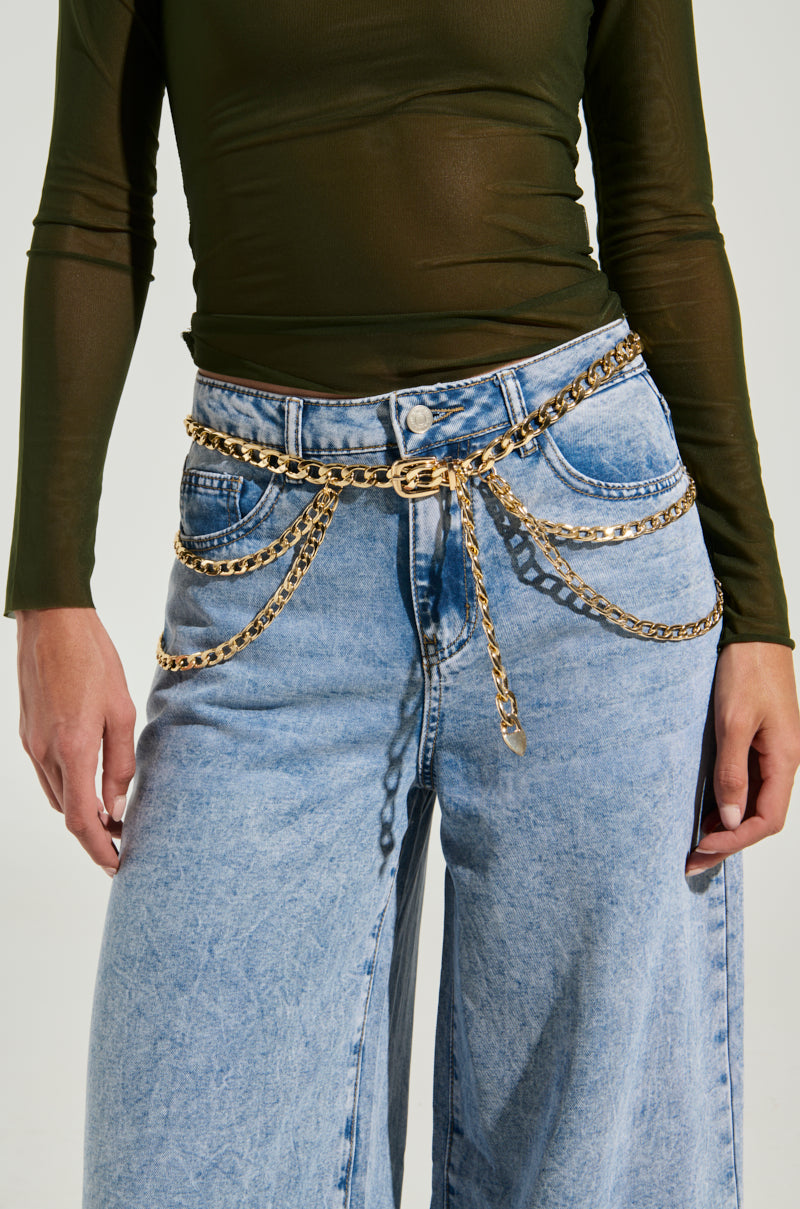 A person wearing light blue, high-waisted, wide-leg jeans and a dark green, long-sleeved, semi-sheer top. The jeans are accessorized with the WESTERN LAYERED CHAIN BELT that drapes across the waist in multiple layers. The background is plain and light-colored. 1