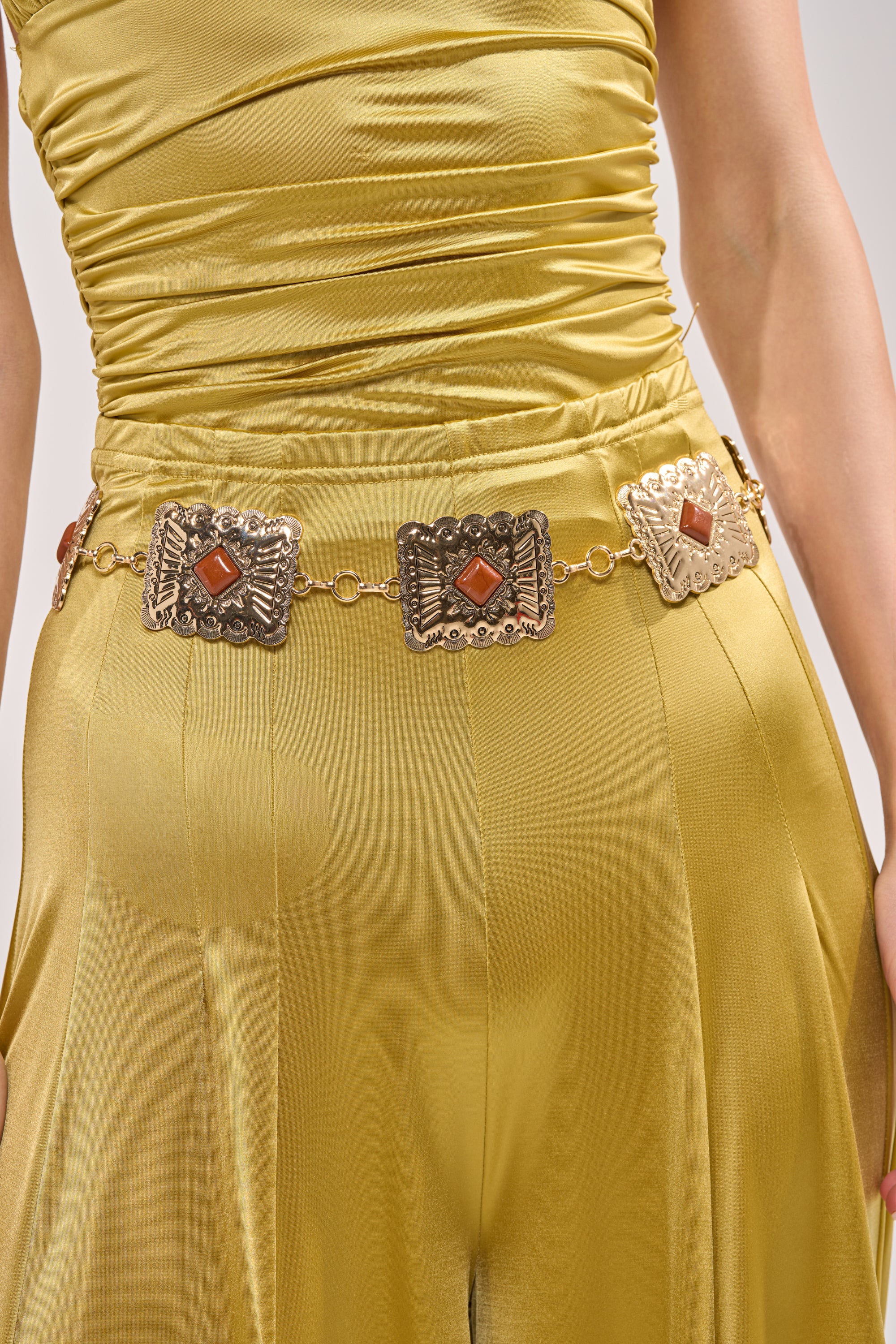 A person wears a shiny yellow dress with the LOCKED IN LOVE CHAIN BELT—a decorative gold chain belt featuring three square plates, each with an orange-red gem. The image highlights their waist and hips. 0