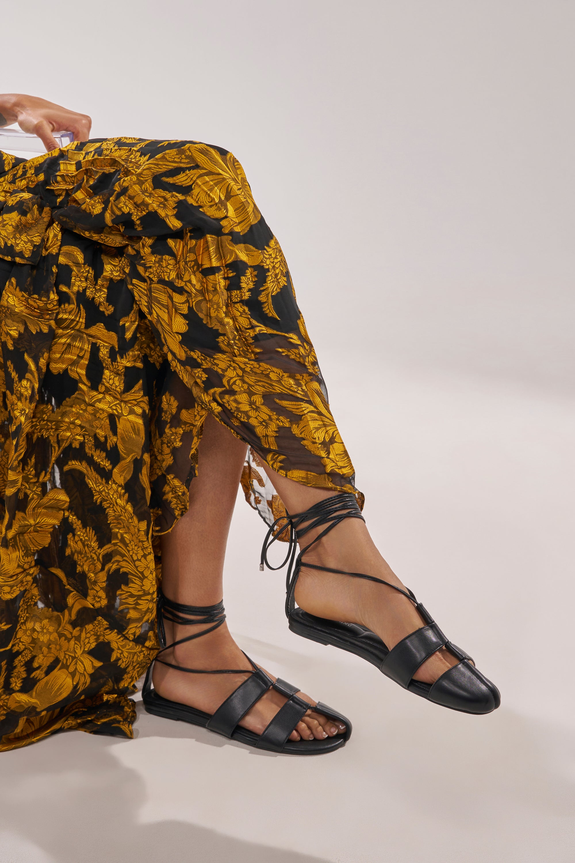 Wearing the AZALEA WANG ICHIRO BLACK PEEP TOE FLAT, a person in a flowing black and yellow floral skirt sits on a light background, one leg casually crossed over the other. 2