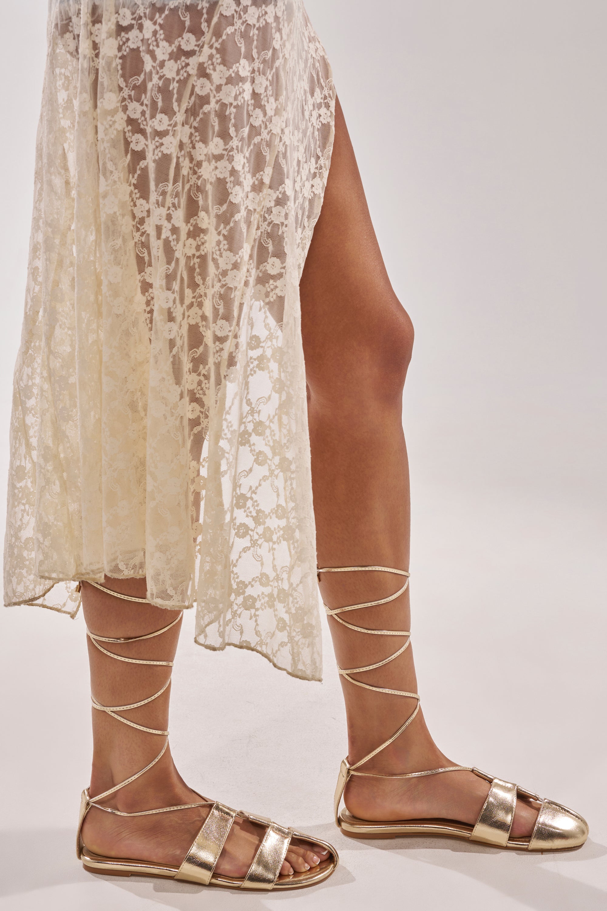 Someone wearing the AZALEA WANG ICHIRO GOLD PEEP TOE FLAT with metallic lace-up design, comfort cushion insole, and a sheer cream lace high-slit skirt, stands against a light background. 1
