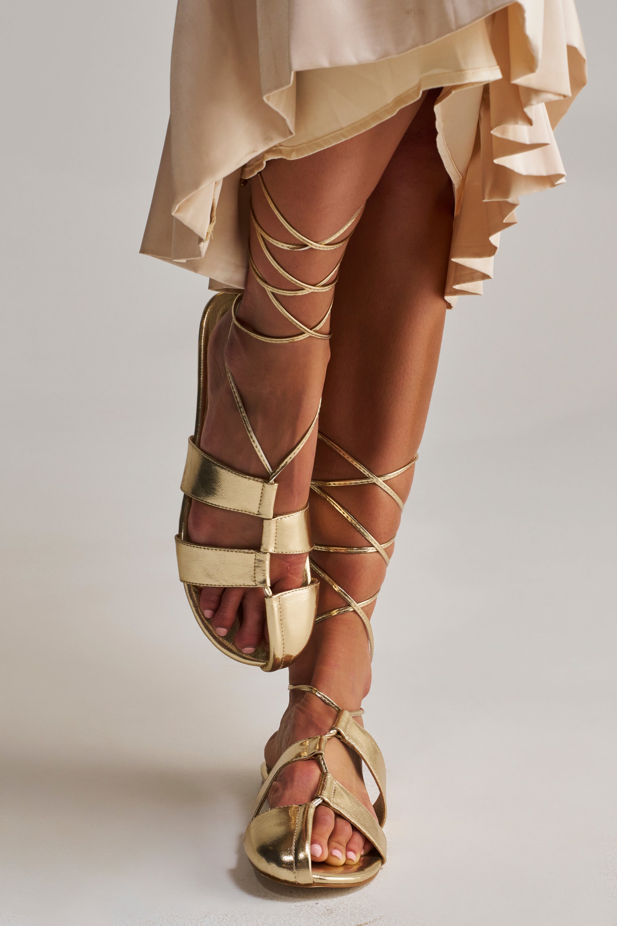 A person wears a light, ruffled skirt and AZALEA WANG ICHIRO GOLD PEEP TOE FLAT sandals with long straps wrapped around their calves, standing on a neutral background.