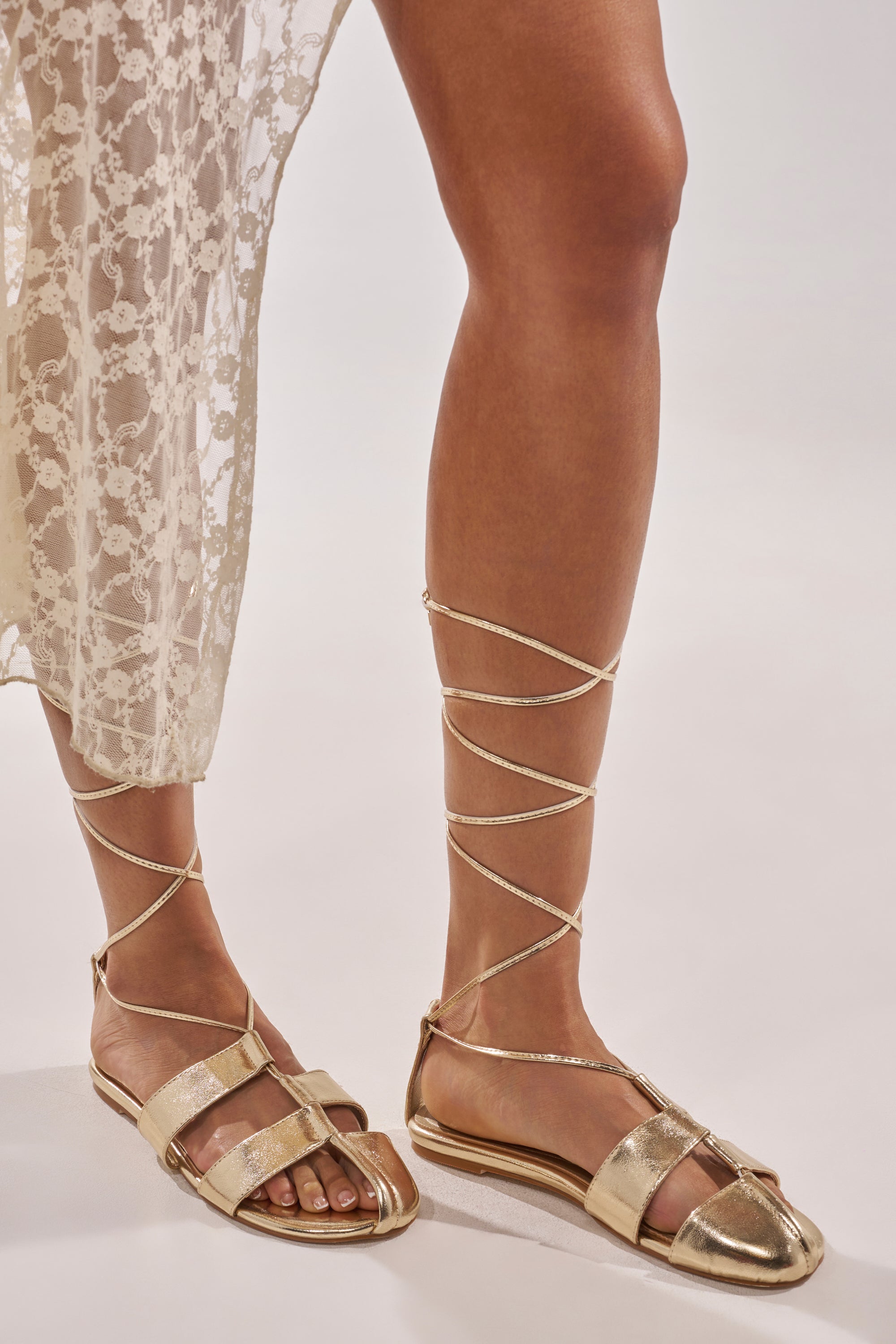 Wearing a sheer lace dress and the AZALEA WANG ICHIRO GOLD PEEP TOE FLAT sandals with comfort cushion insoles, featuring lace-up straps wrapped around the lower legs, a person stands against a plain light background. 3