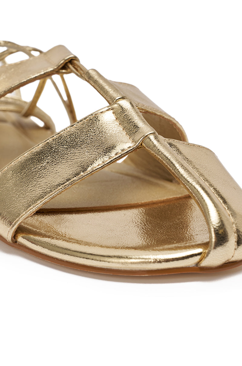Close-up of the AZALEA WANG ICHIRO GOLD PEEP TOE FLAT, showcasing shiny gold metallic crisscross straps, an open toe, and a comfort cushion insole atop a smooth, light brown sole. 7