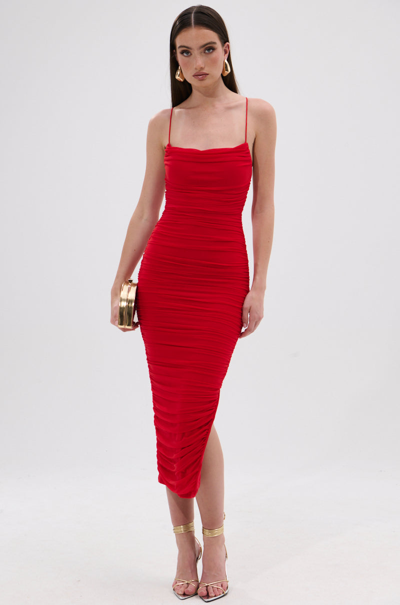 A woman stands against a white background wearing the SHINE BRIGHT SLINKY MIDI DRESS in red, styled with gold hoop earrings, strappy gold heels, and a gold clutch. 0