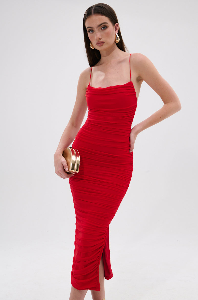 Wearing the SHINE BRIGHT SLINKY MIDI DRESS, a woman with straight brown hair and gold hoop earrings stands confidently against a plain background, holding a small gold clutch. The fitted bodycon dress features ruched details and spaghetti straps. 1