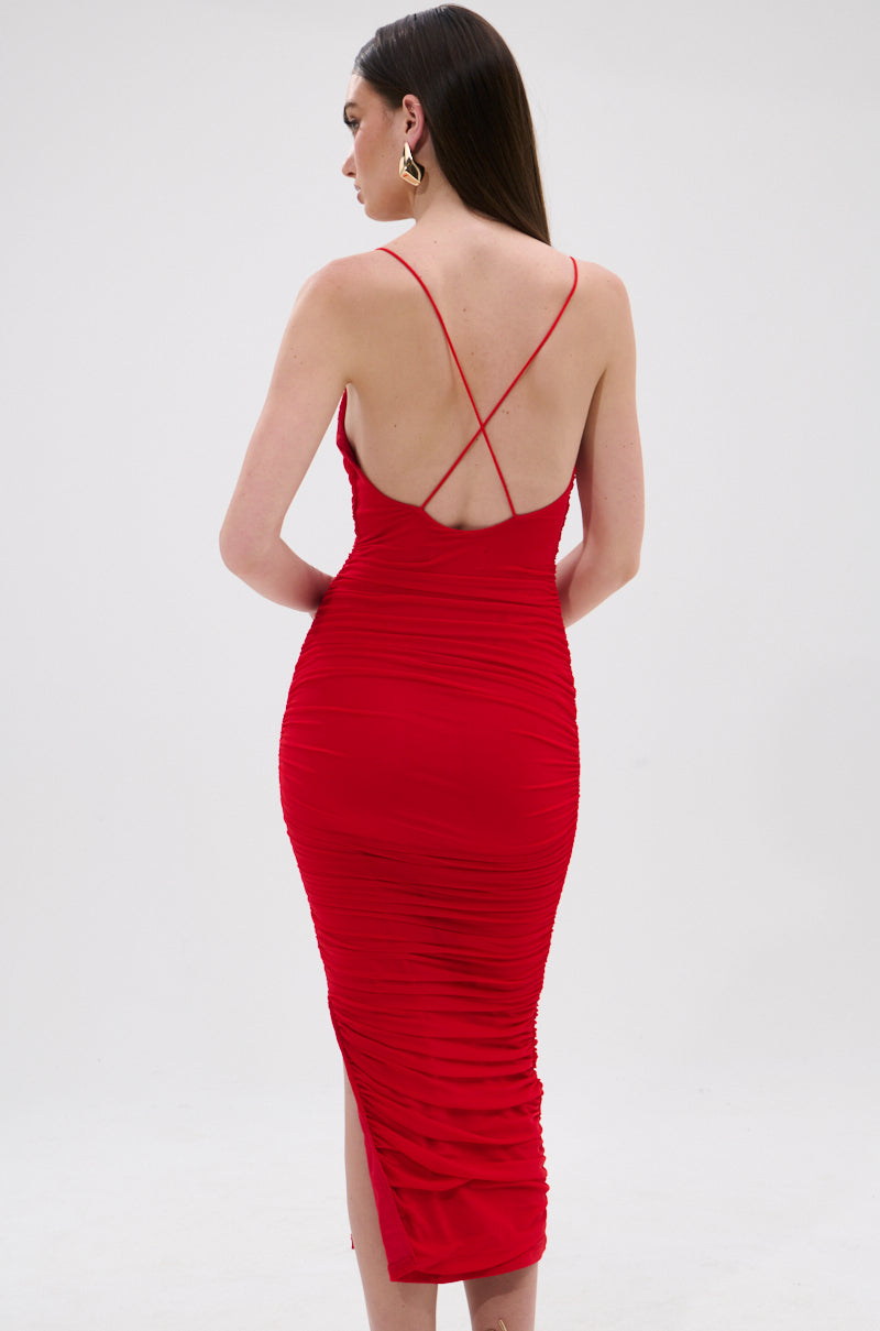 A woman stands with her back to the camera against a plain light background, wearing the SHINE BRIGHT SLINKY MIDI DRESS—a fitted red bodycon midi dress with thin criss-cross straps and ruched detailing for a flattering silhouette. 3