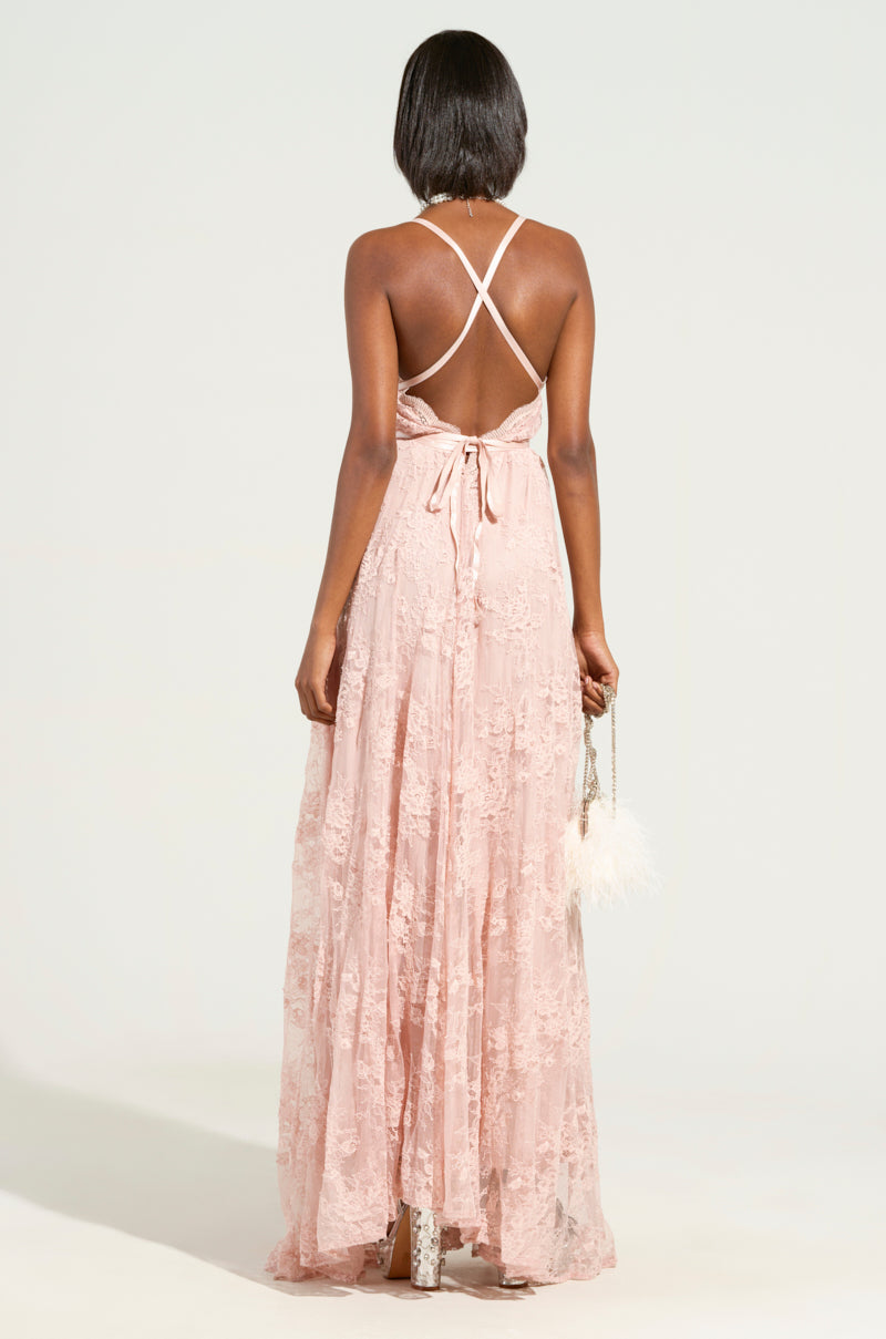 Dressed in the stunning CRAVE YOU LACE MAXI DRESS, featuring a pink, semi-stretchy lace fabric with an open back and crisscross straps, a woman holds a small feather-adorned purse. Her high heels complete the ensemble, while the plunging v-neckline adds allure against the simple, light-colored background. 4