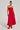 A woman is posing in the SITTING PRETTY POPLIN MIDI DRESS IN RED, a strapless, ankle-length dress featuring a ruched bodice and a flared skirt. She complements her outfit with red shoes and an elegant updo hairstyle. The background is plain and white.