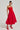 A woman stands in a minimalistic room, wearing the eye-catching SITTING PRETTY POPLIN MIDI DRESS IN RED, featuring a ruched bodice and an A-line skirt. She has her hair styled up and is wearing red, strappy high-heeled shoes, exuding confidence and poise.