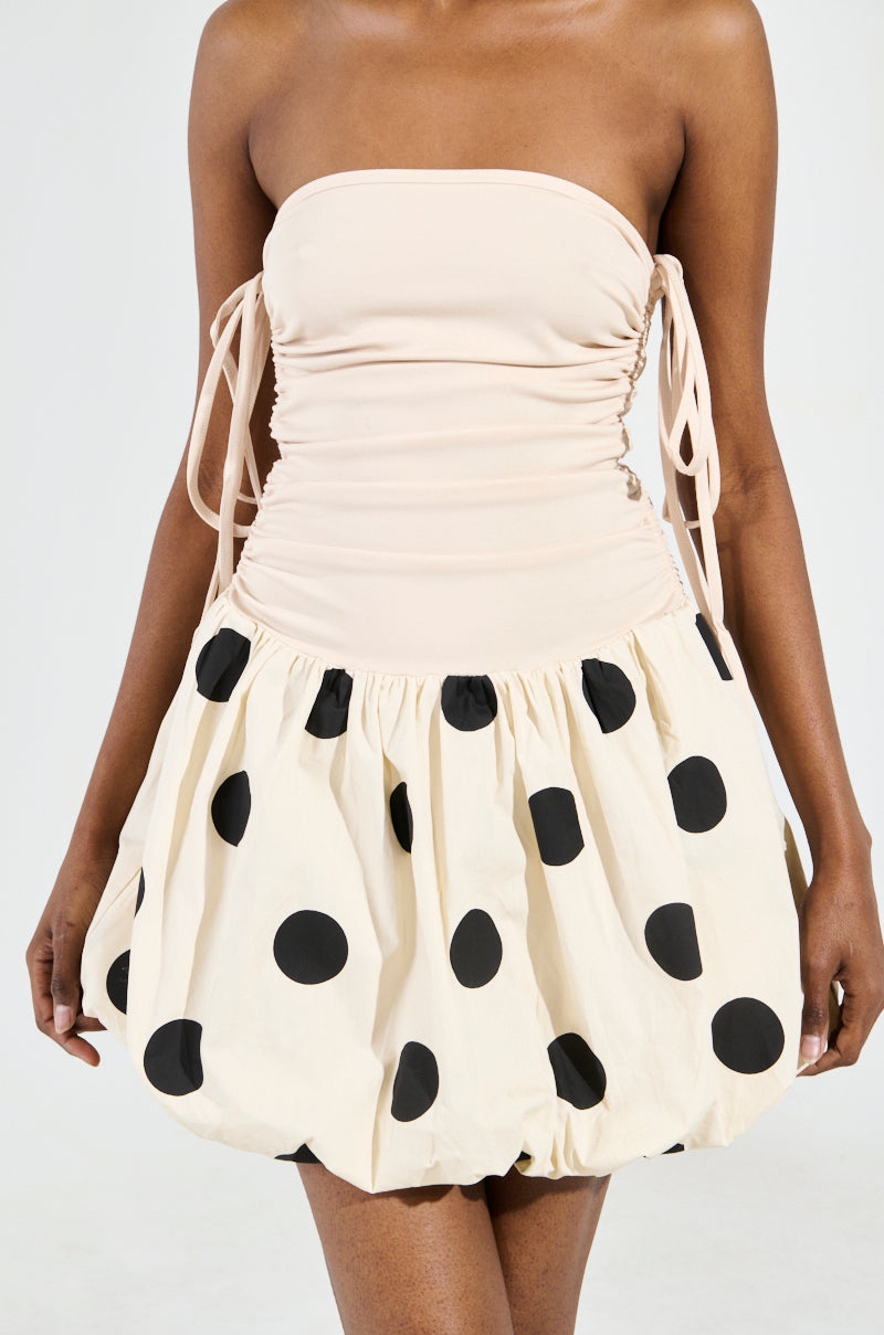 A person is wearing the ON TREND BUBBLE MINI DRESS, which showcases a beige ruched top and a cream puffy skirt with an eye-catching bubble hem decorated with large black polka dots. The dress also features tied side details, as the person stands against a plain light background.
