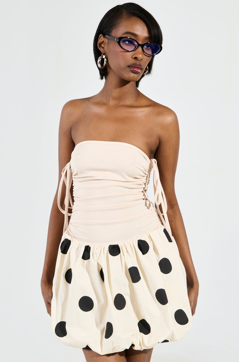 A person is wearing the ON TREND BUBBLE MINI DRESS, featuring a fitted beige top and a puffed skirt with large black polka dots. They have short hair, accessorized with blue-tinted sunglasses and silver hoop earrings, standing against a plain background.