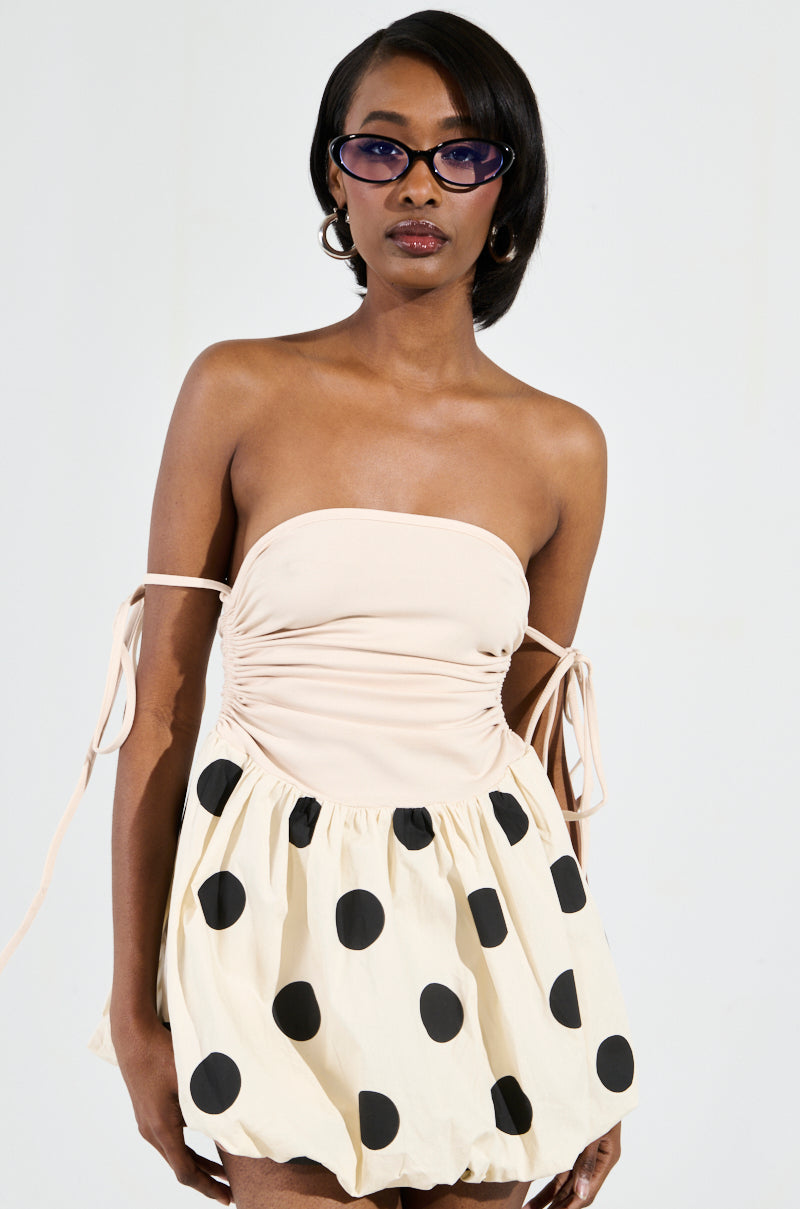 A person with short dark hair wears purple sunglasses and the ON TREND BUBBLE MINI DRESS, showcasing a polka dot pattern with large black spots and off-the-shoulder ties. The background is a plain, light color.
