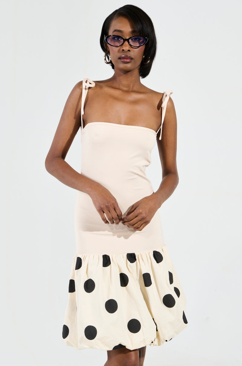 A woman in the ON TREND BUBBLE MINI DRESS, featuring a beige hue and a striking polka dot pattern on the skirt, stands against a plain background. She stylishly poses with sunglasses and her hands together in front of her.