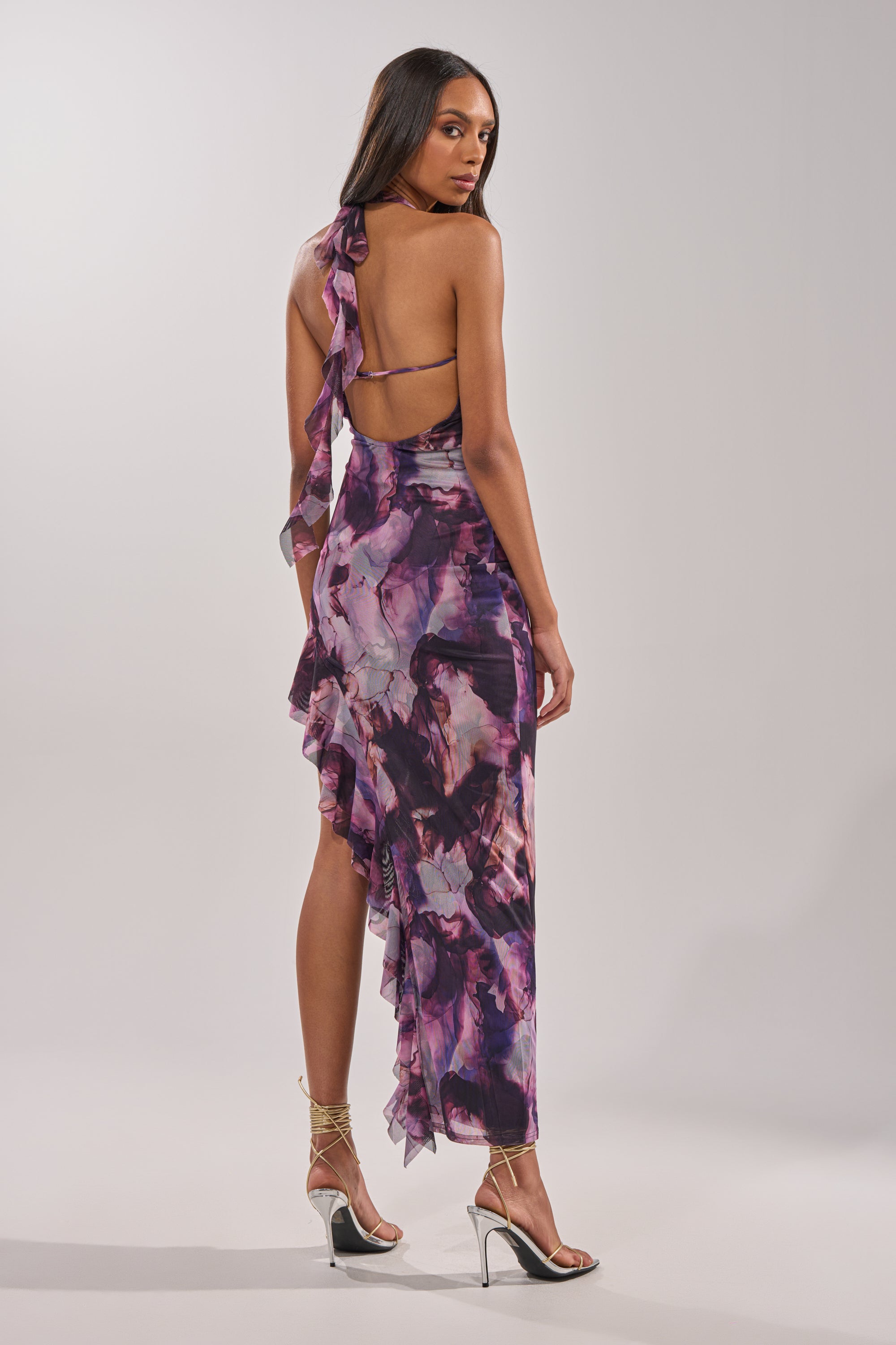 A woman poses in the BAECATION MIDI DRESS, a sleeveless purple floral midi with ruffles and an open back. She wears silver strappy heels, facing away over her shoulder against a plain background—ideal for vacation. 1