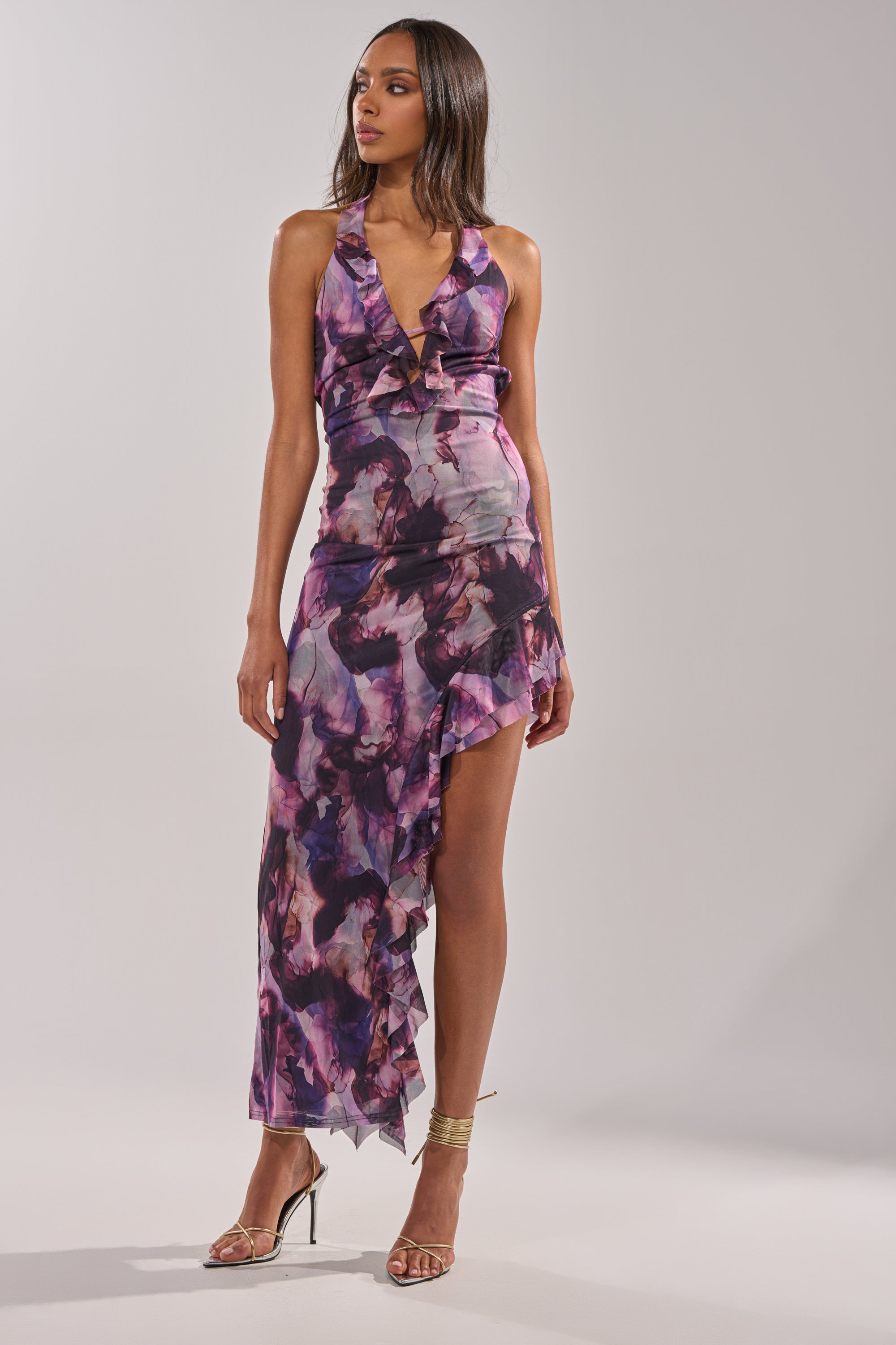 A woman wears the BAECATION MIDI DRESS, a sleeveless purple and black floral midi with a deep V-neck and asymmetric ruffled hem, paired with gold strappy high-heeled sandals against a plain light background. 2
