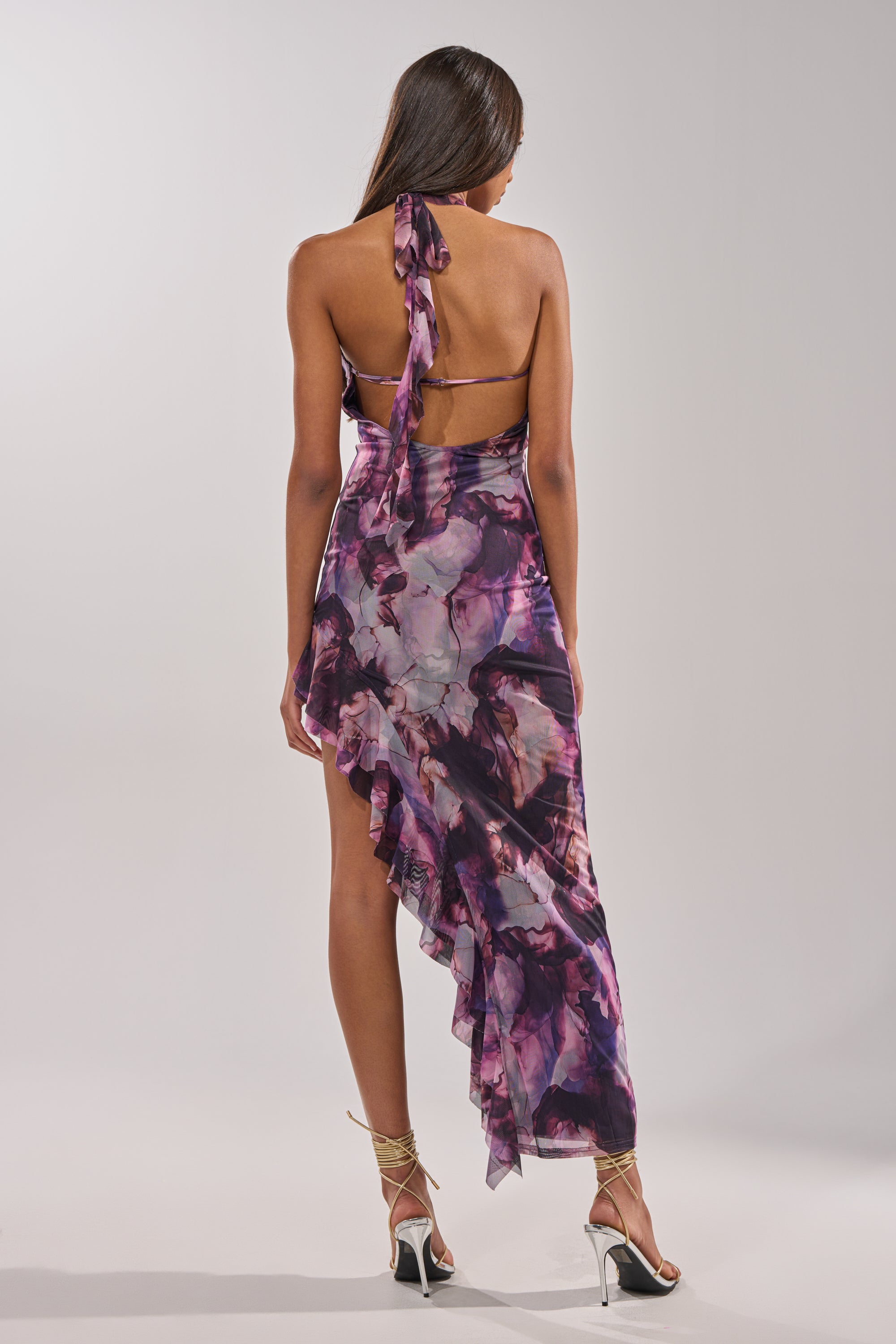 A woman faces away, wearing the BAECATION MIDI DRESS, a halter neck style with a purple and pink marble pattern and ruffled hem—ideal for vacations. She completes her effortlessly chic look with strappy high-heeled sandals. 5
