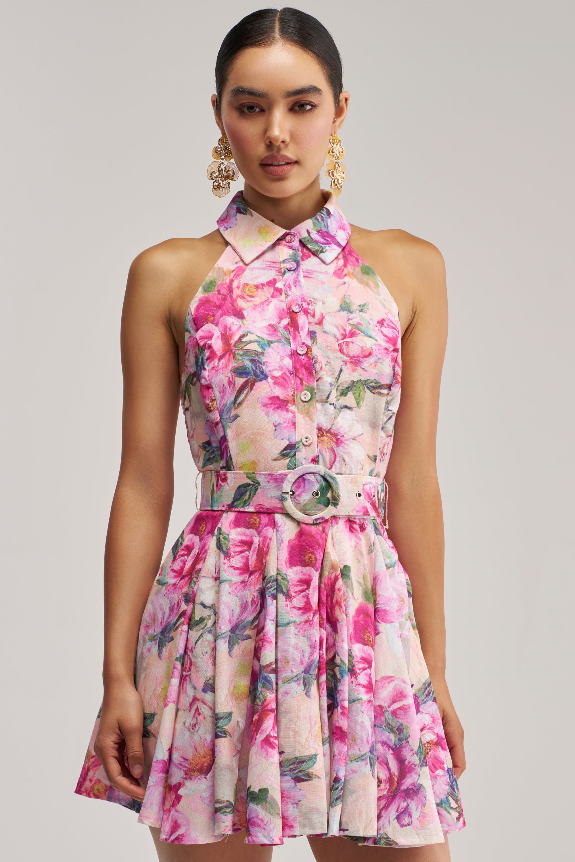 A woman stands against a plain background wearing the SHES A DIVA FLORAL MINI DRESS IN PINK MULTI—a sleeveless, collared mini dress with a pink floral print, matching belt, and gold statement earrings.
