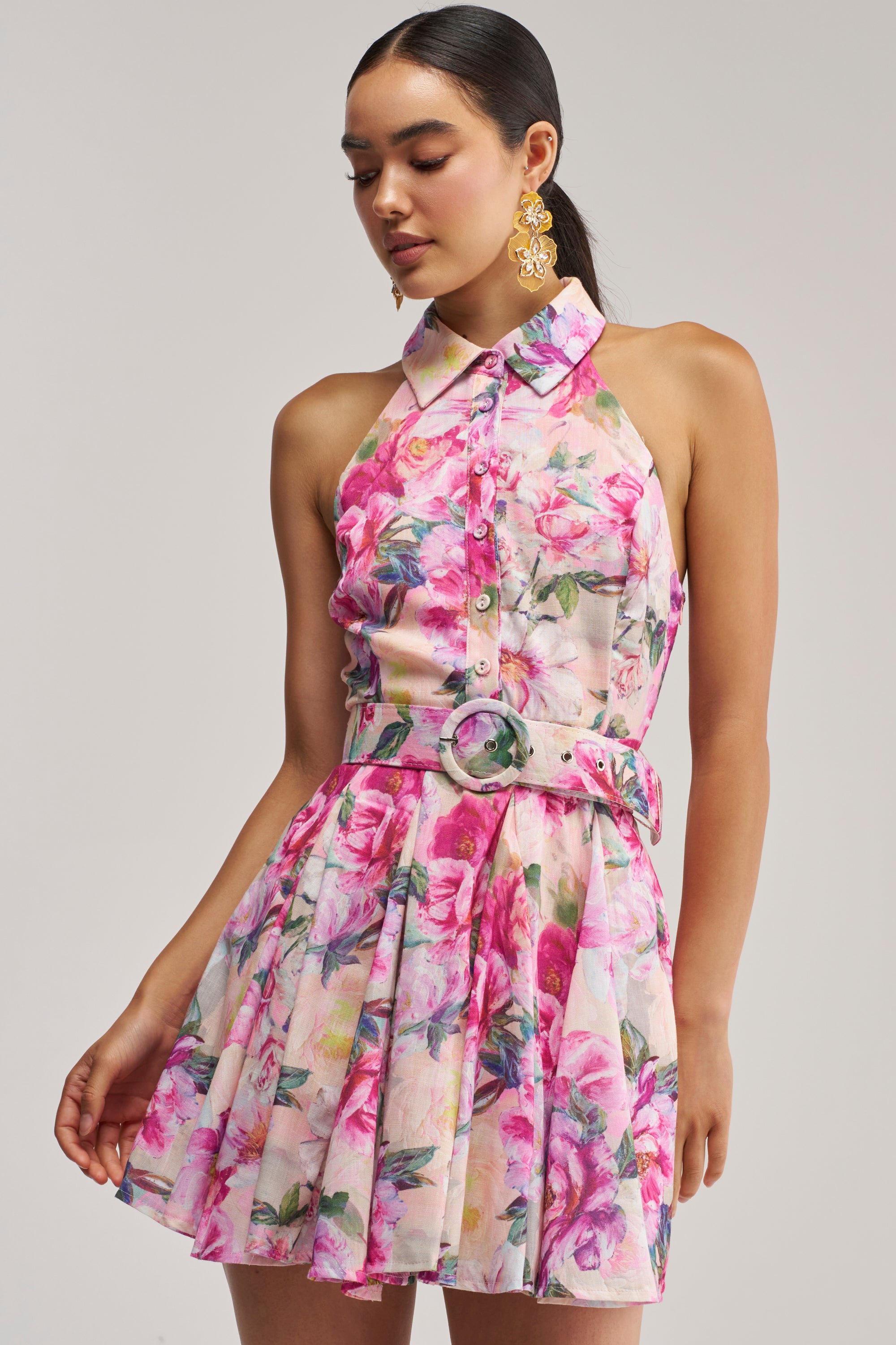 A woman models the SHES A DIVA FLORAL MINI DRESS IN PINK MULTI—a sleeveless, collared pink floral mini dress with a matching belt—paired with gold statement earrings and styled with a sleek low ponytail, standing against a plain light gray background.