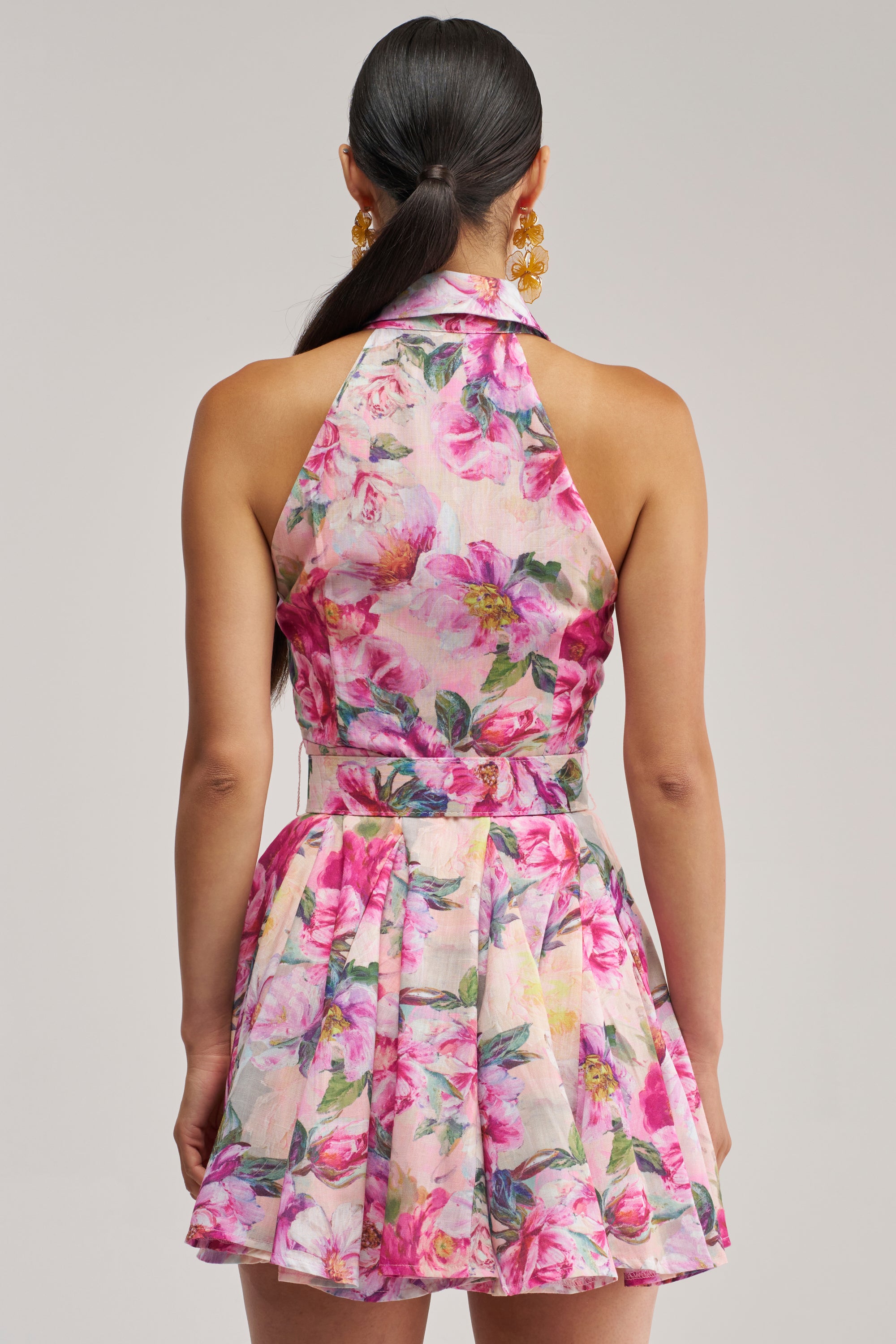 A woman with dark hair in a ponytail is seen from the back, wearing the SHES A DIVA FLORAL MINI DRESS IN PINK MULTI, a sleeveless mini dress featuring a belted waist and delicate pink and white rose patterns. She accessorizes with large gold flower-shaped earrings against a neutral background.