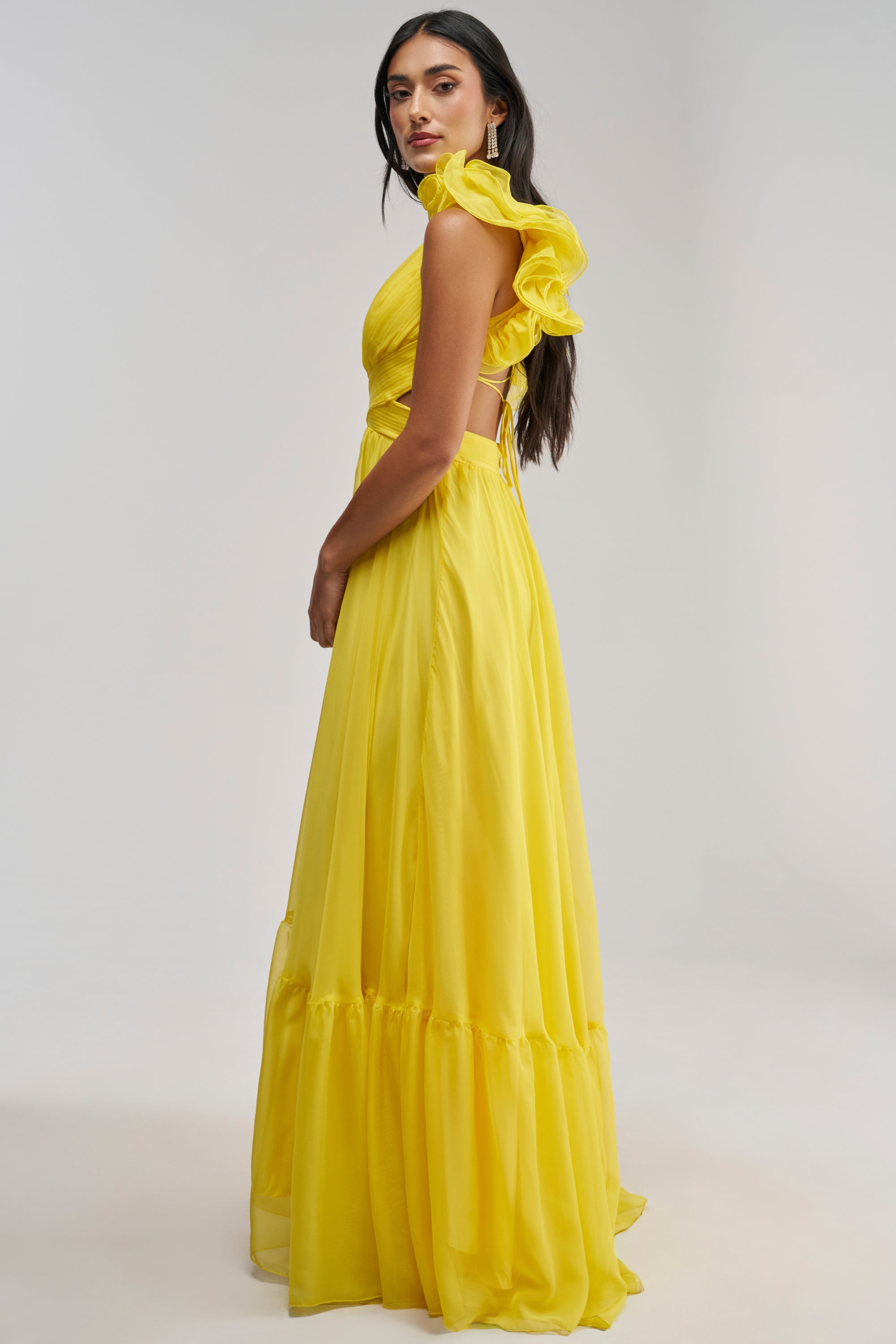 A woman stands sideways against a plain light background, wearing the POPPING CHAMPAGNE CHIFFON MAXI DRESS IN YELLOW. The flowing chiffon maxi features ruffled sleeves and vibrant color, beautifully complemented by her long dark hair and calm expression—making it an ideal choice for a summer wedding guest dress.