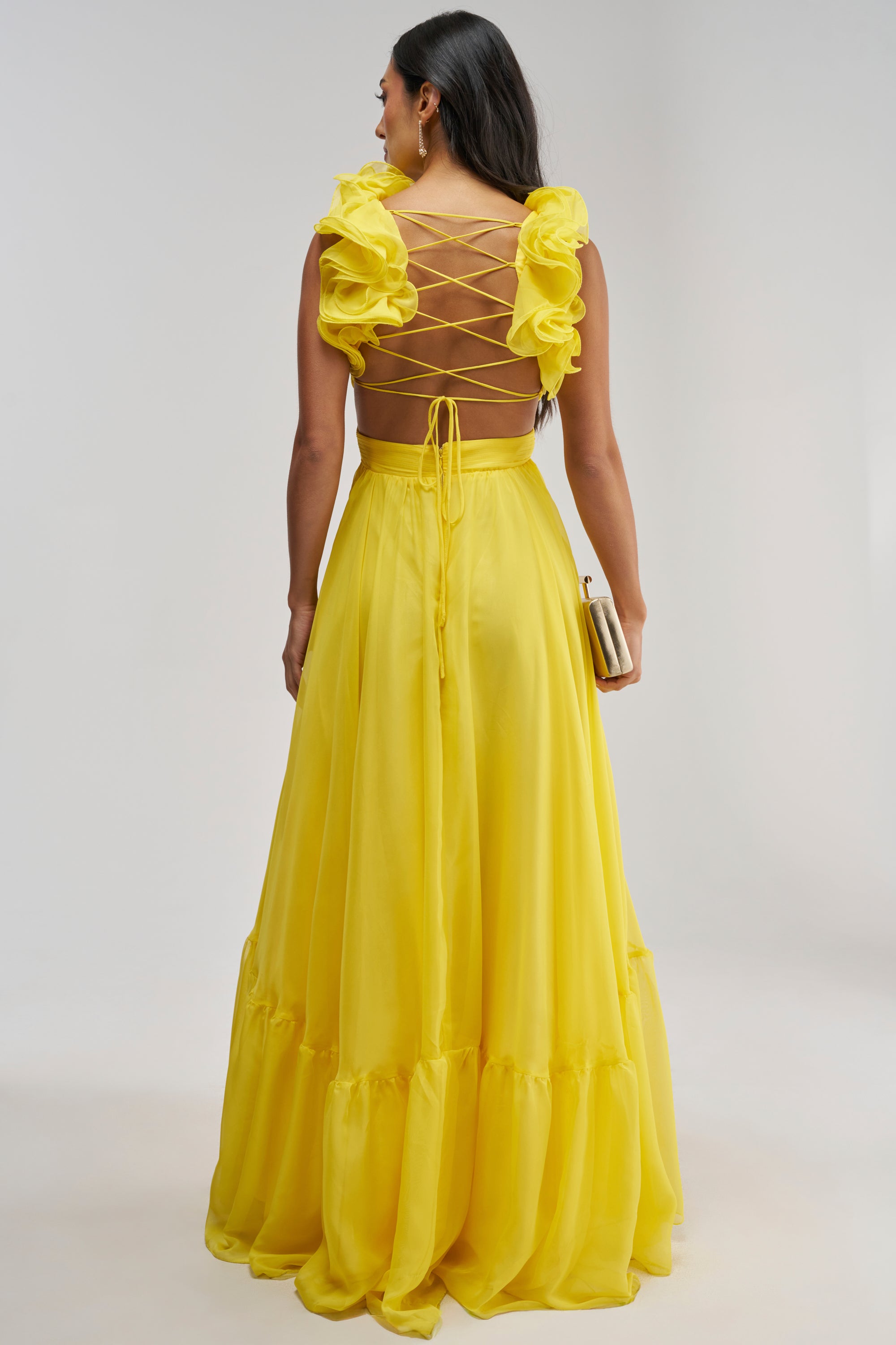 Dressed in the POPPING CHAMPAGNE CHIFFON MAXI DRESS IN YELLOW, a woman stands against a plain light background, the flowing ruffled sleeves and lace-up back making this the perfect summer wedding guest dress—she holds a small clutch to complete her elegant look.