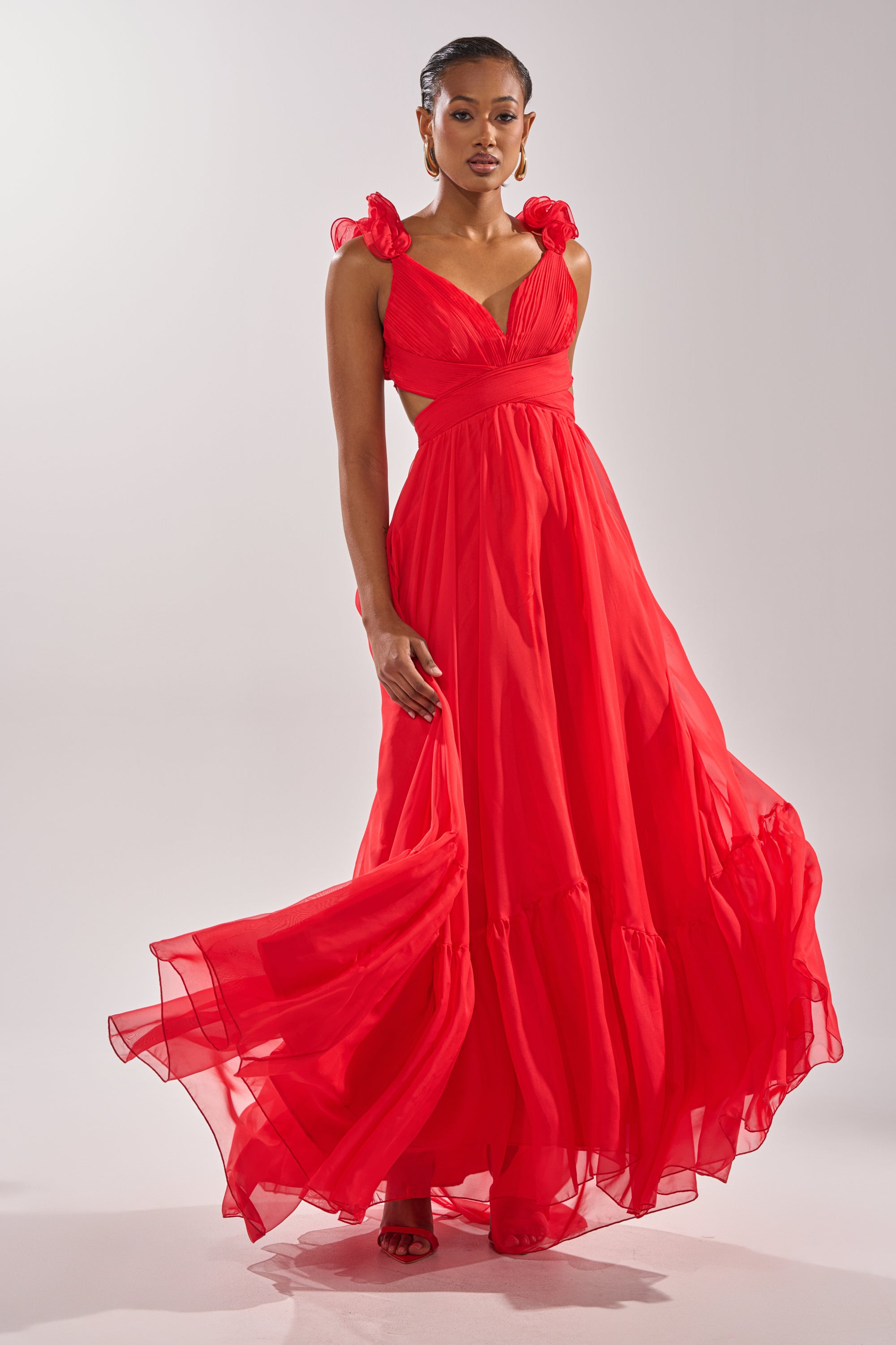 A woman in the DREAM GIRL MAXI DRESS IN RED, featuring ruffle shoulder details and light, airy fabric, stands confidently against a white background, highlighting the dress’s elegant, flowing full-length silhouette. 0