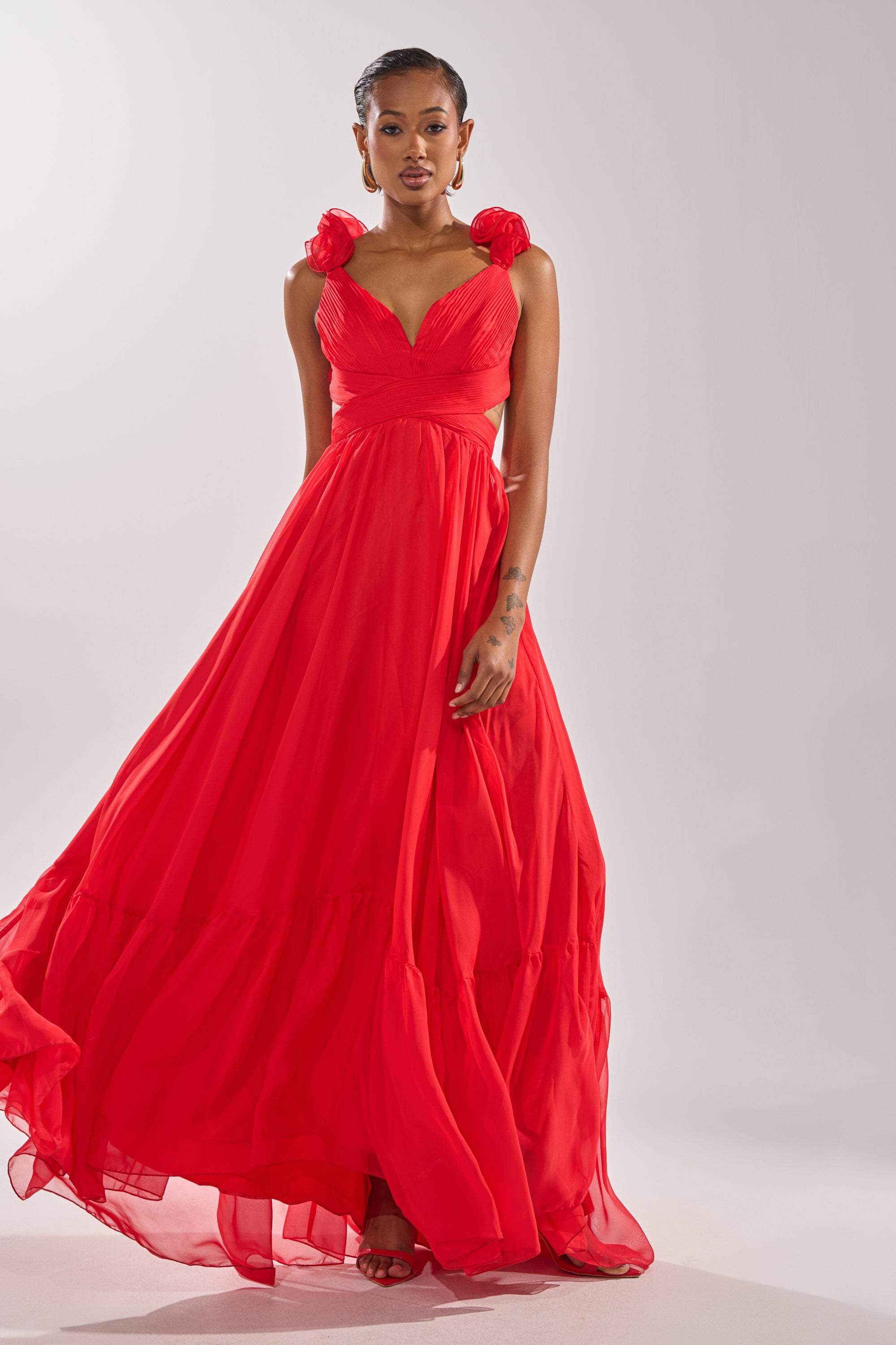 A woman wears the DREAM GIRL MAXI DRESS IN RED, featuring floral shoulder details, a fitted bodice, and a corseted lace-up back—an elegant choice for any special occasion—against a plain light background. 1