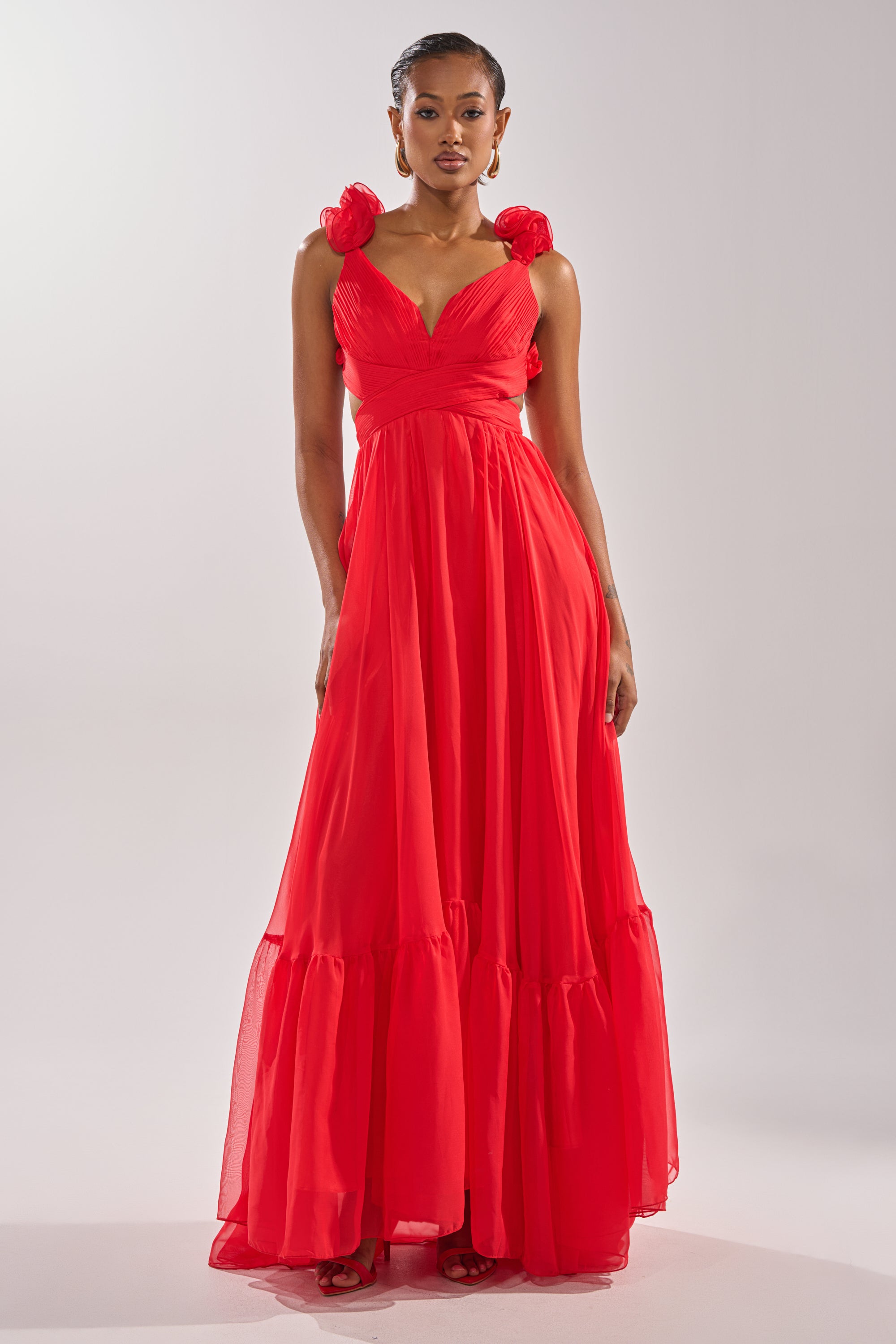 A woman models the DREAM GIRL MAXI DRESS IN RED, featuring ruffle shoulder details, a v-neckline, and large flower accents on the straps, set against a plain light background. 3