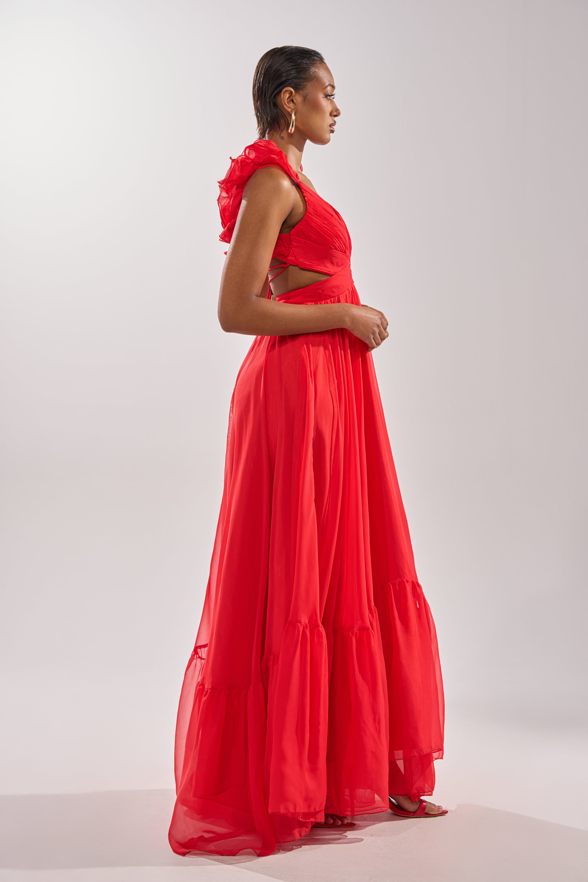 A woman stands in profile against a plain light background, wearing the DREAM GIRL MAXI DRESS IN RED featuring ruffle shoulder details and waist cut-outs. 4