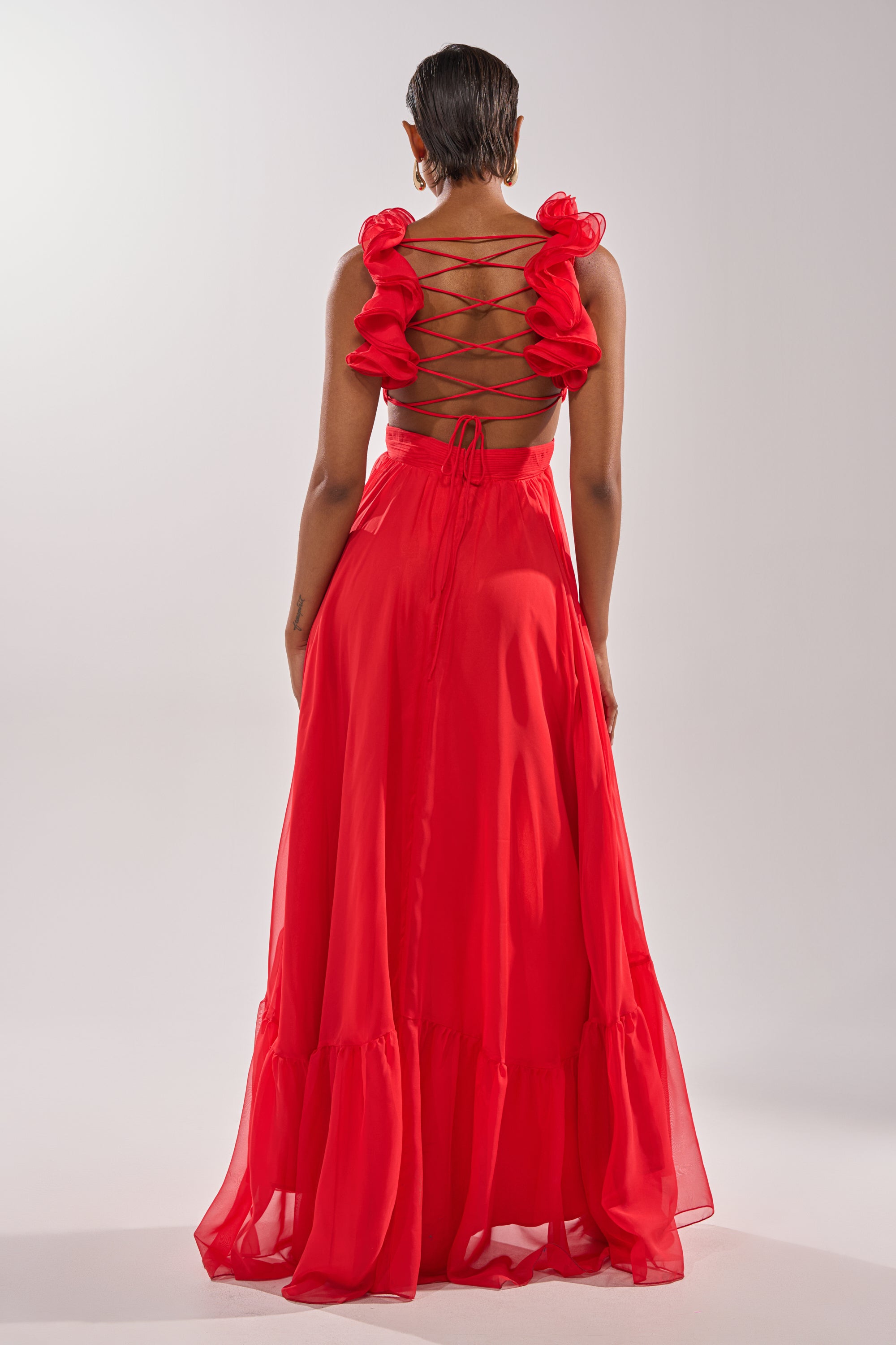 A person models the DREAM GIRL MAXI DRESS IN RED, featuring ruffled shoulder details and a corset back, standing with their back to the camera against a plain light background. 5