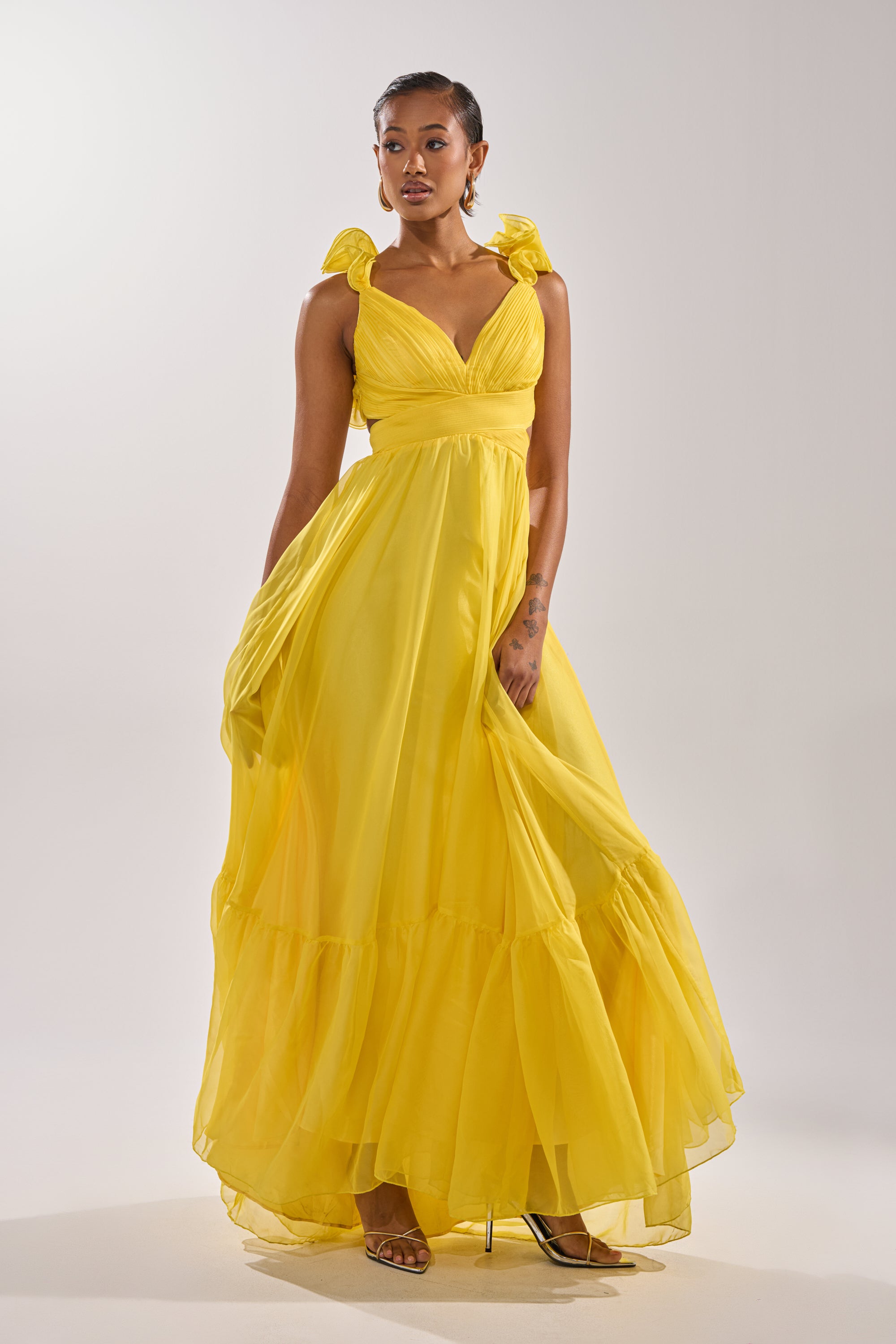 A woman in the DREAM GIRL MAXI DRESS IN YELLOW with ruffled shoulder straps and a tiered ruffle hem stands gracefully, holding her skirt. She has short hair, hoop earrings, and wears strappy heels.