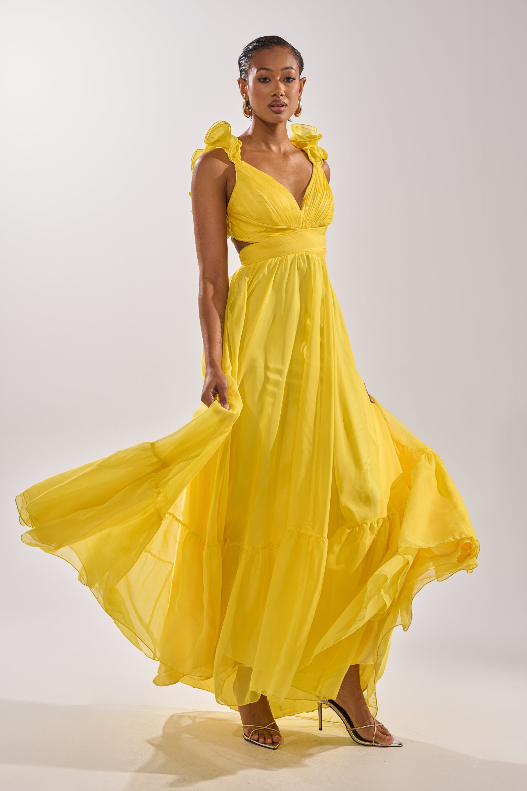 A woman poses against a plain light background wearing the DREAM GIRL MAXI DRESS IN YELLOW, a flowing maxi dress with ruffled shoulder details and strappy high heels. The dress billows out gracefully as she stands.