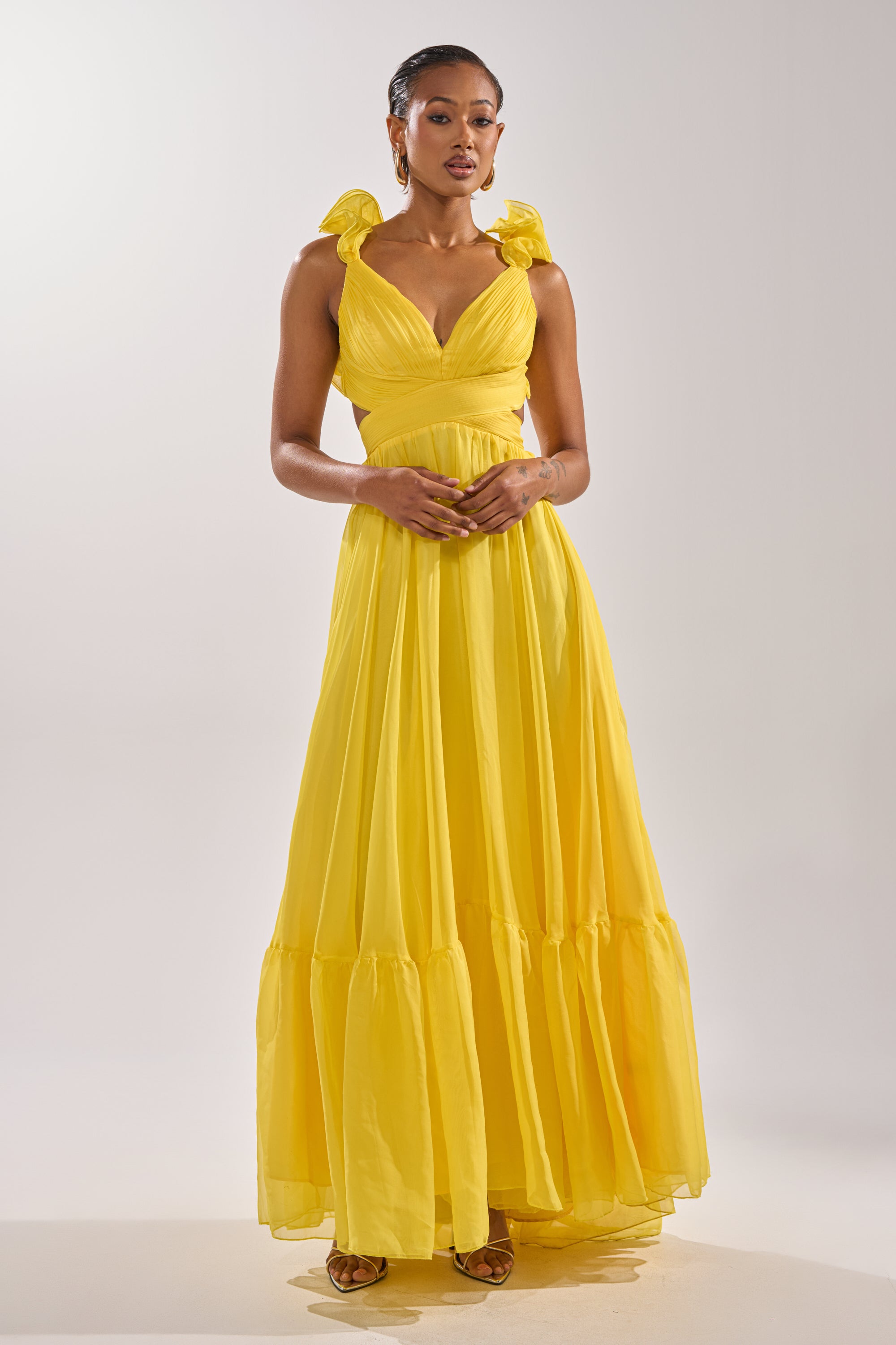 A woman poses against a plain, light background wearing the DREAM GIRL MAXI DRESS IN YELLOW, featuring ruffle shoulder straps and a flowing skirt. She gently clasps her hands in front of her.