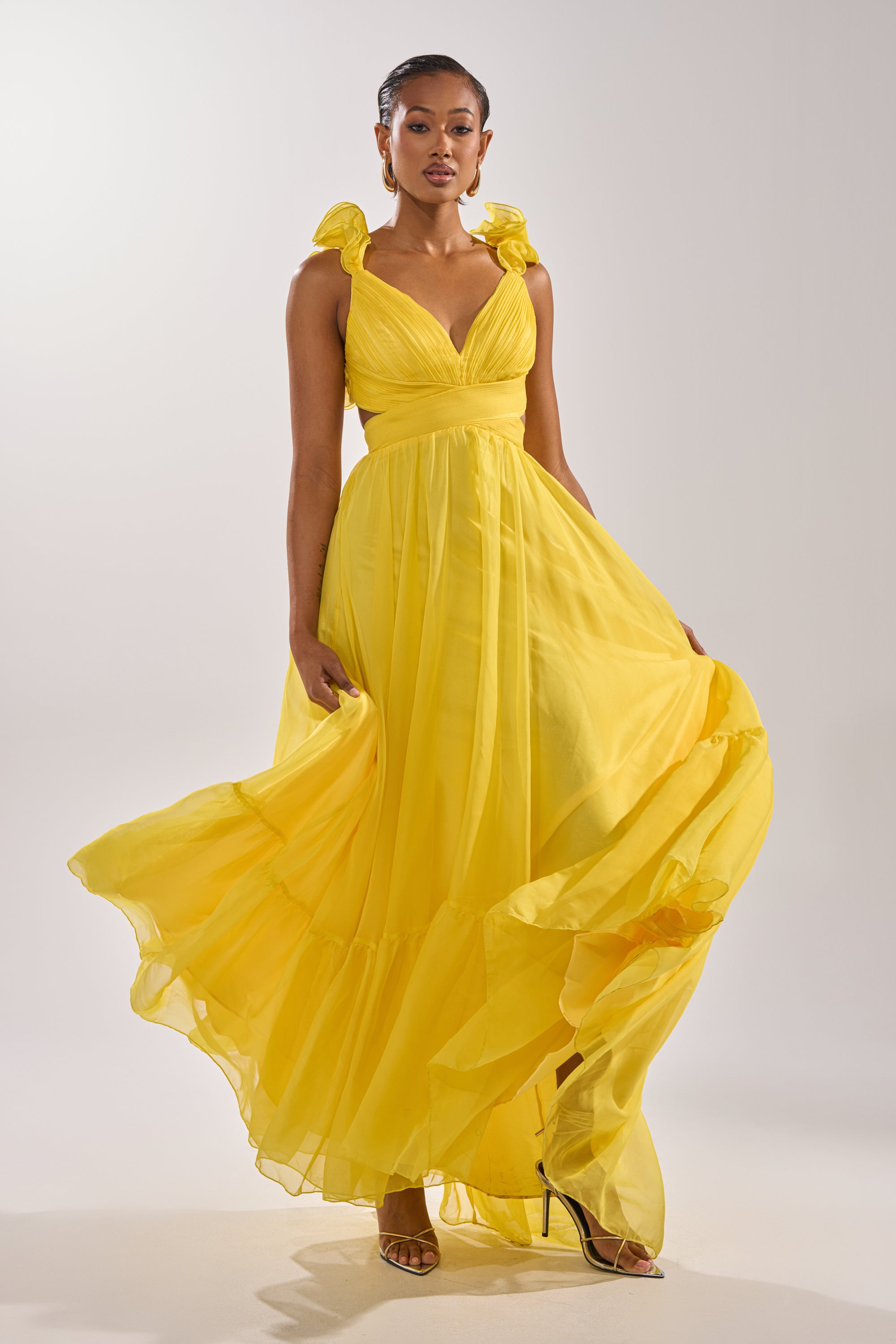 A woman poses confidently against a plain background, elegantly holding the skirt of the DREAM GIRL MAXI DRESS IN YELLOW, which features a bright hue and ruffle hem details.