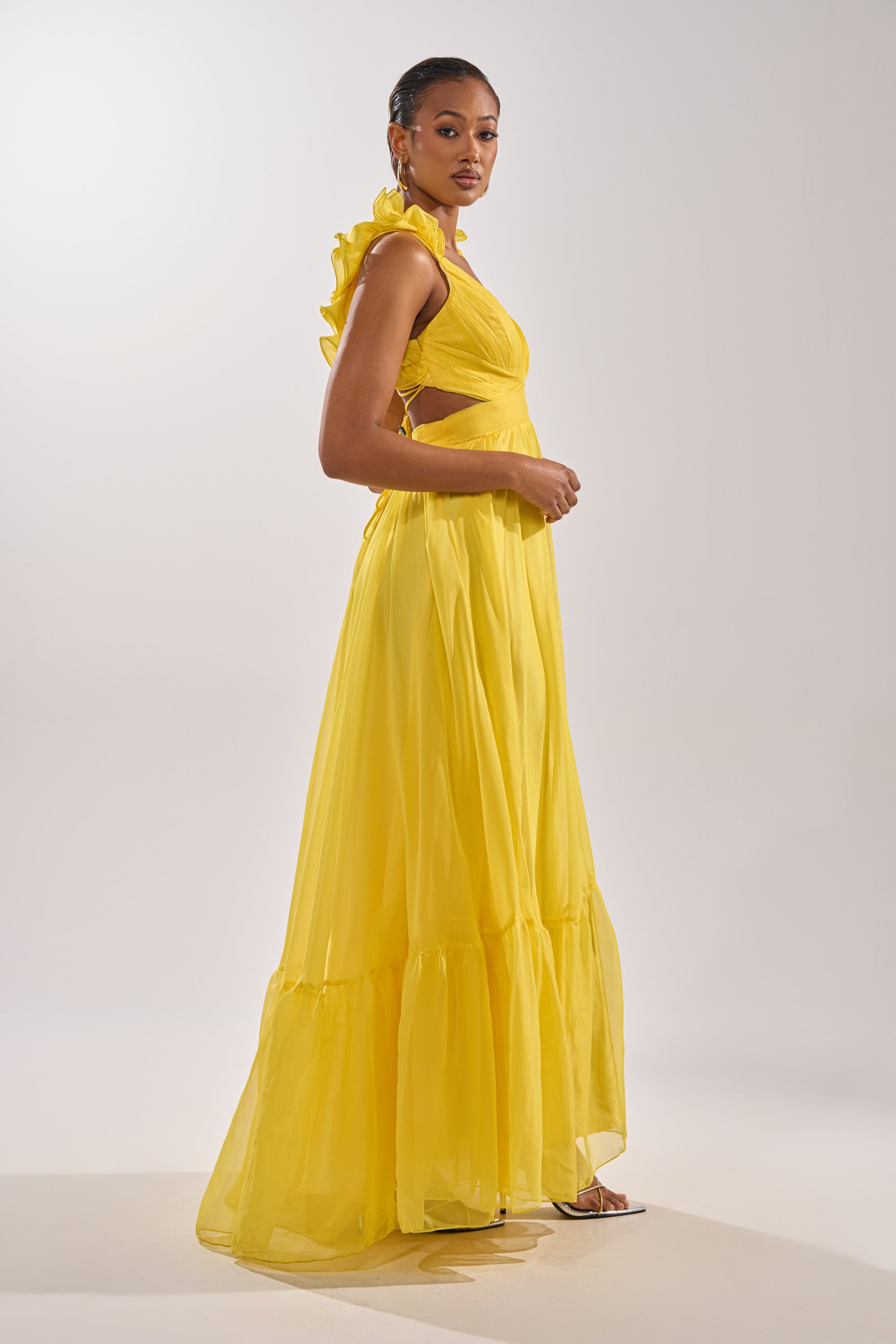 A woman stands in profile wearing the DREAM GIRL MAXI DRESS IN YELLOW, featuring a ruffle hem and lace-up back. The simple, light background highlights her elegant, flowing look.