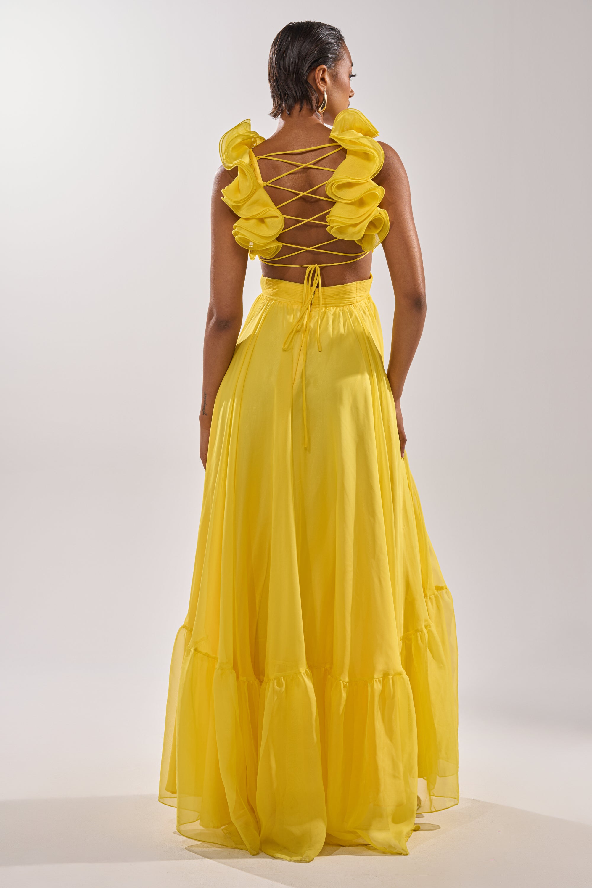 A woman faces away, wearing the DREAM GIRL MAXI DRESS IN YELLOW with a ruffle hem and lace-up back. The flowing skirt stands out against a plain, light backdrop for a fresh, striking look.