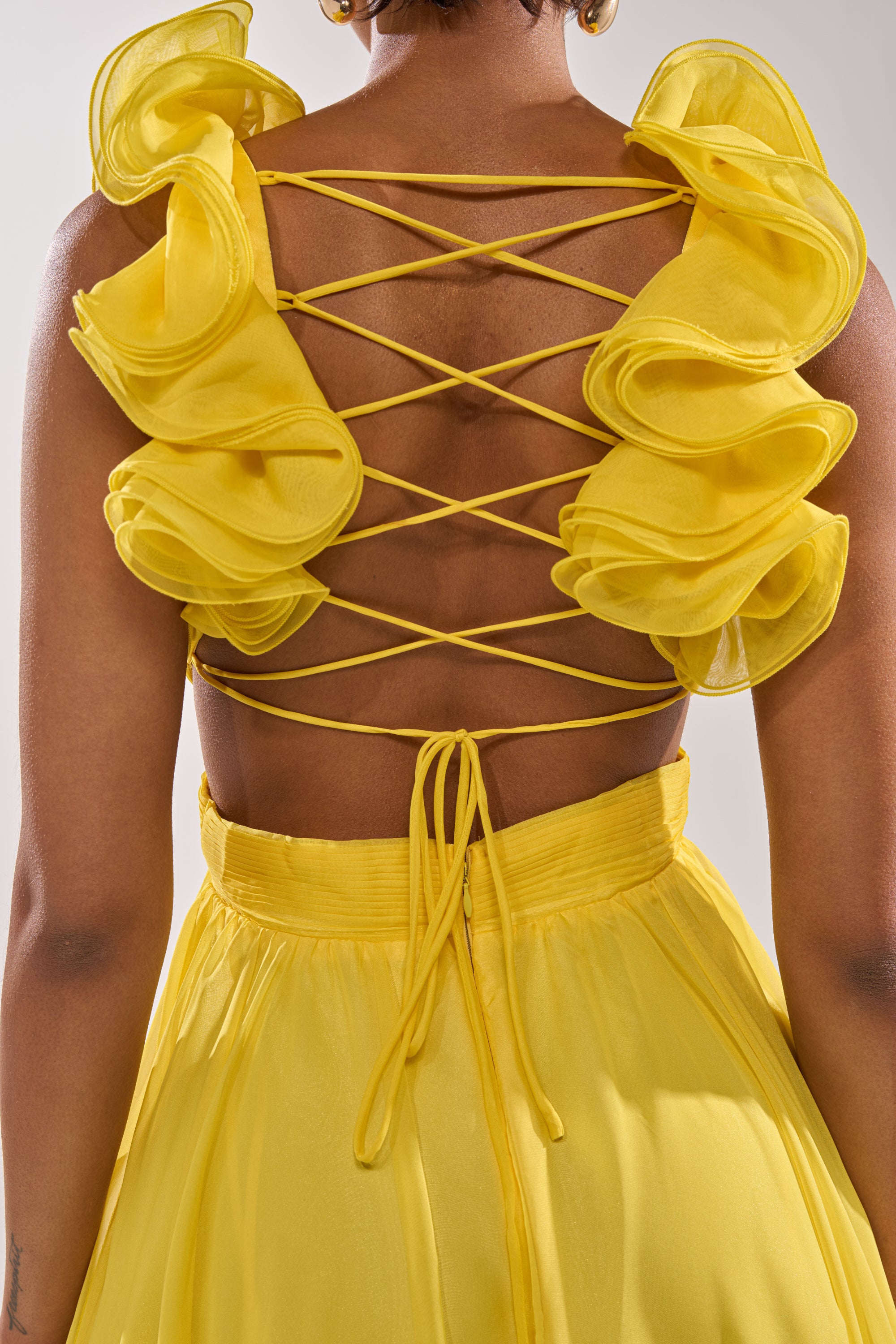 A person wearing the DREAM GIRL MAXI DRESS IN YELLOW featuring ruffled sleeves, a crisscross lace-up back, a ruffled hem, and a waist tie bow.