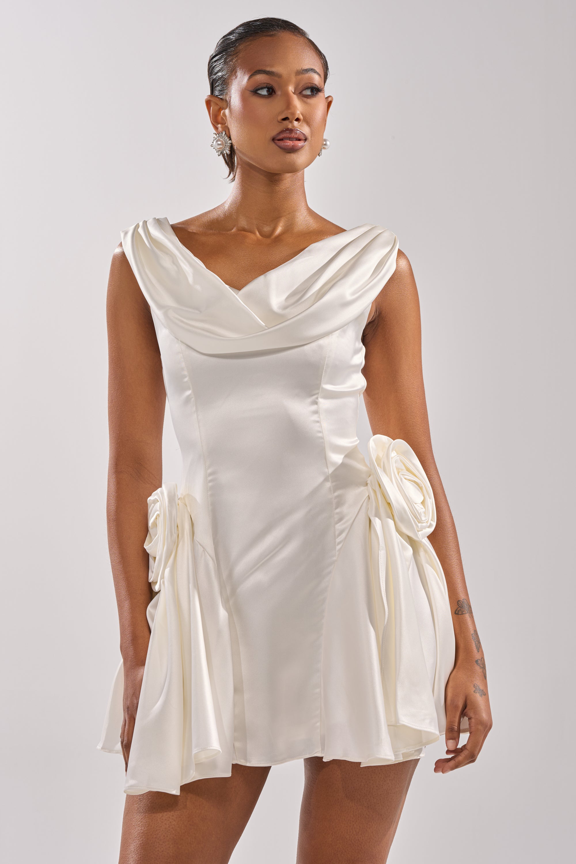 A woman models the SWEET ESCAPE MINI DRESS by AKIRA Label, featuring white satin, a draped neckline, and large fabric flower accents at the hips, posed against a plain light background.