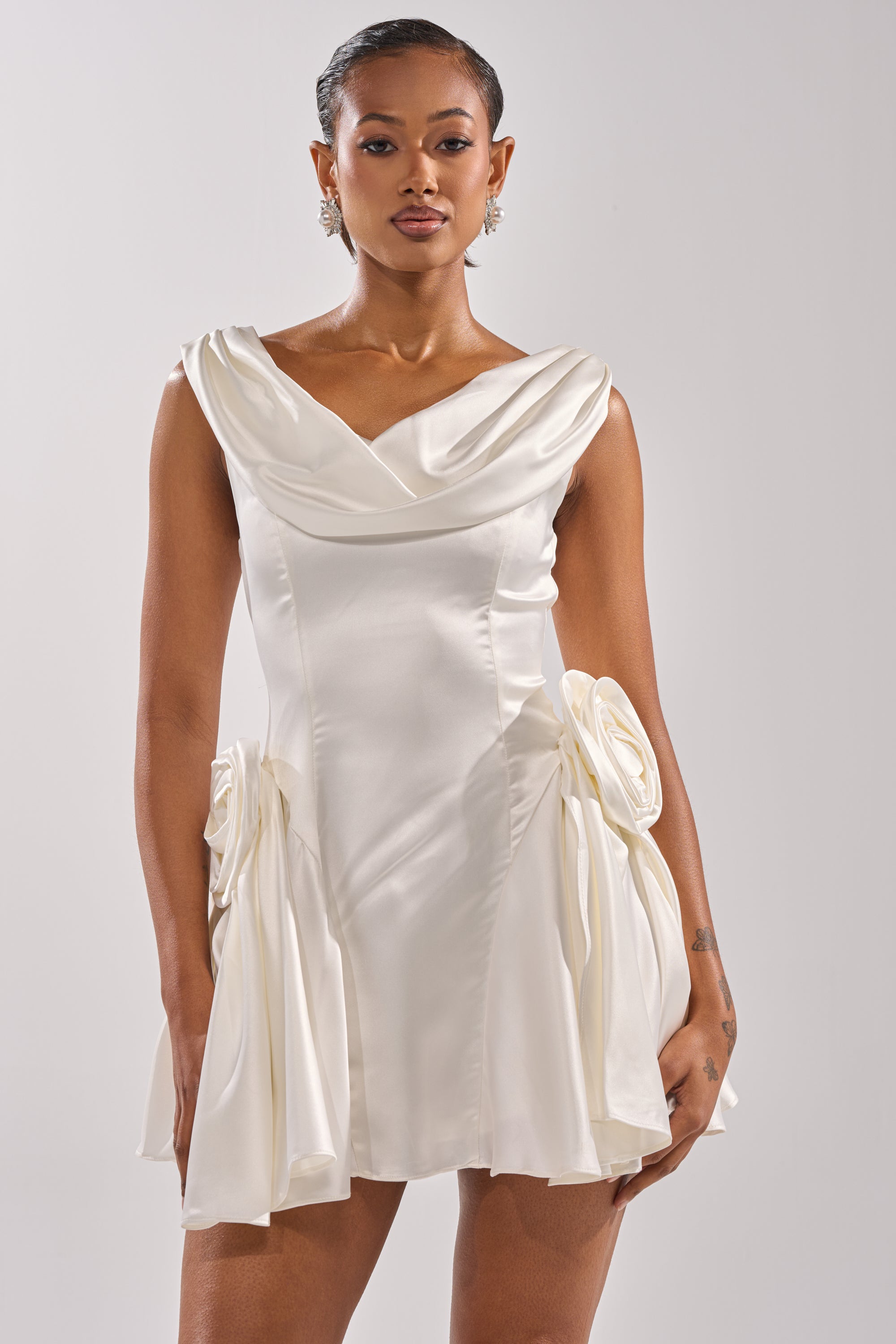 A woman models the SWEET ESCAPE MINI DRESS, a short, sleeveless white satin cocktail dress with a draped neckline and bold fabric accents at the hips, set against a simple light background.