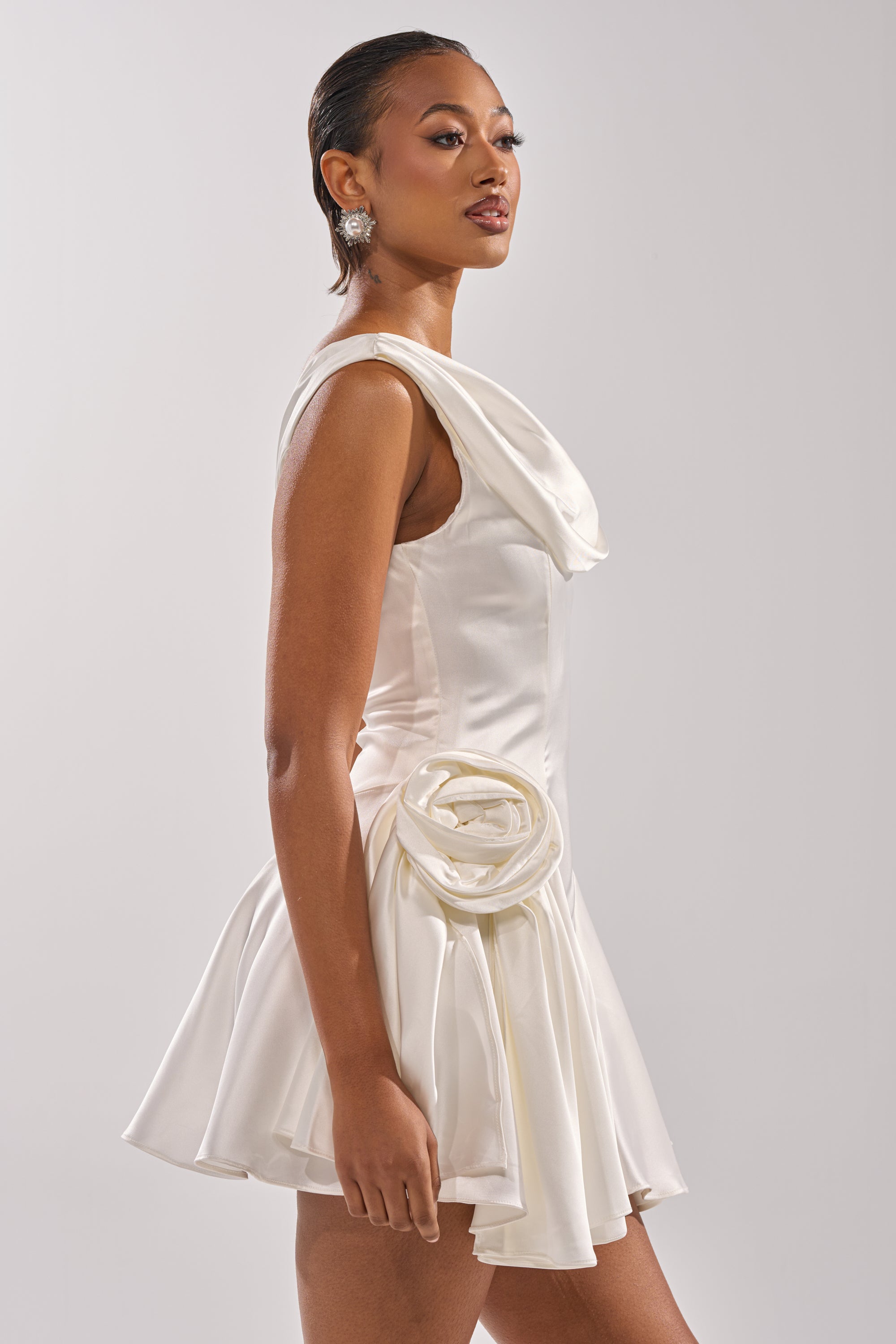 A woman stands sideways against a plain background, wearing the SWEET ESCAPE MINI DRESS—a couture-inspired, sleeveless white satin mini dress featuring a large rosette at the waist. She has slicked-back hair and statement earrings, looking confidently ahead.