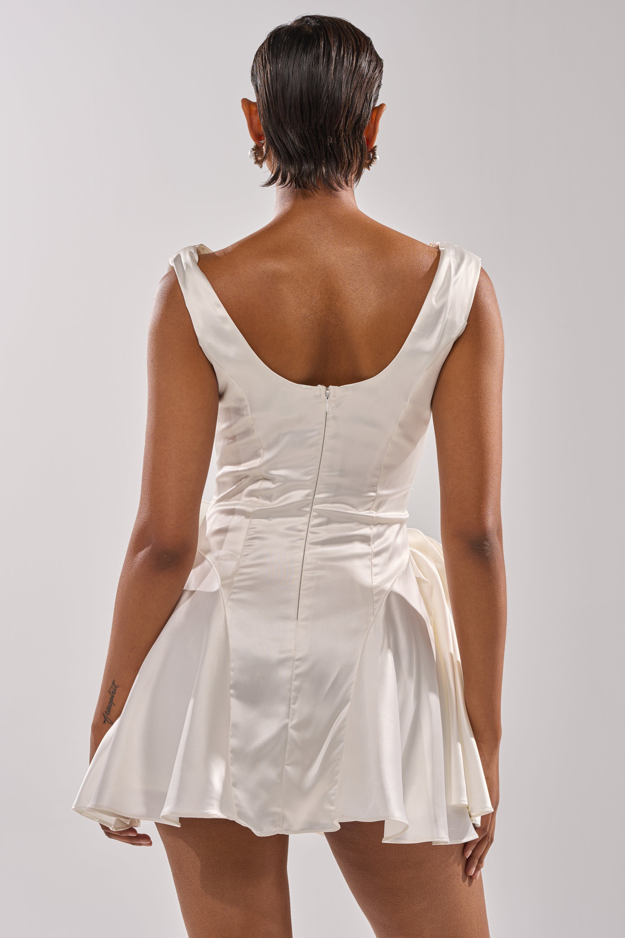 A person with short dark hair is seen from the back wearing the SWEET ESCAPE MINI DRESS, a white satin sleeveless, backless bridal mini dress with a flared skirt and center zipper, set against a plain light background.
