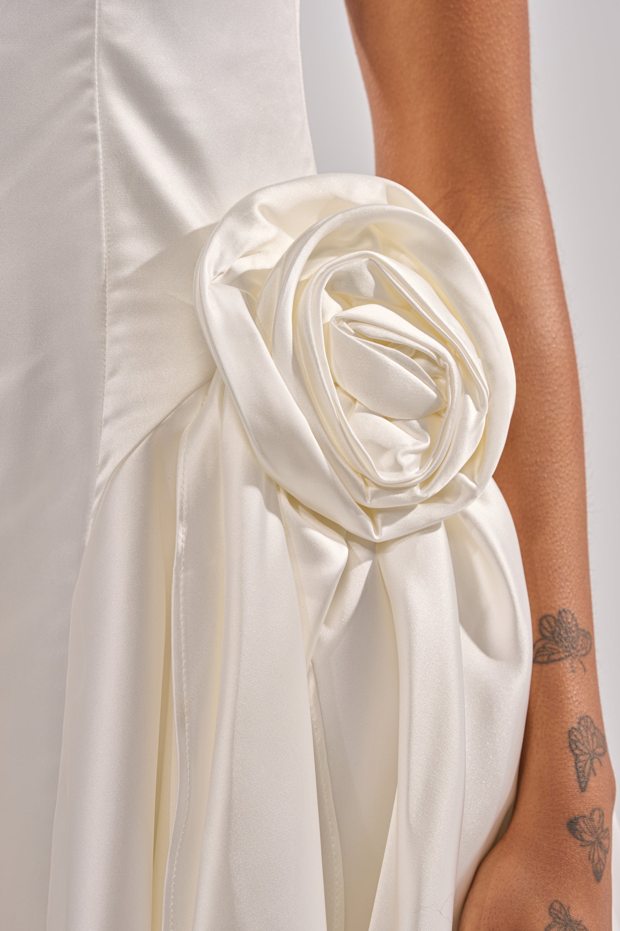 A close-up of the SWEET ESCAPE MINI DRESS, a white satin bridal mini with a large fabric rose at the hip and a cowl neckline. A floral tattoo on a person’s arm is visible beside the decorative flower.