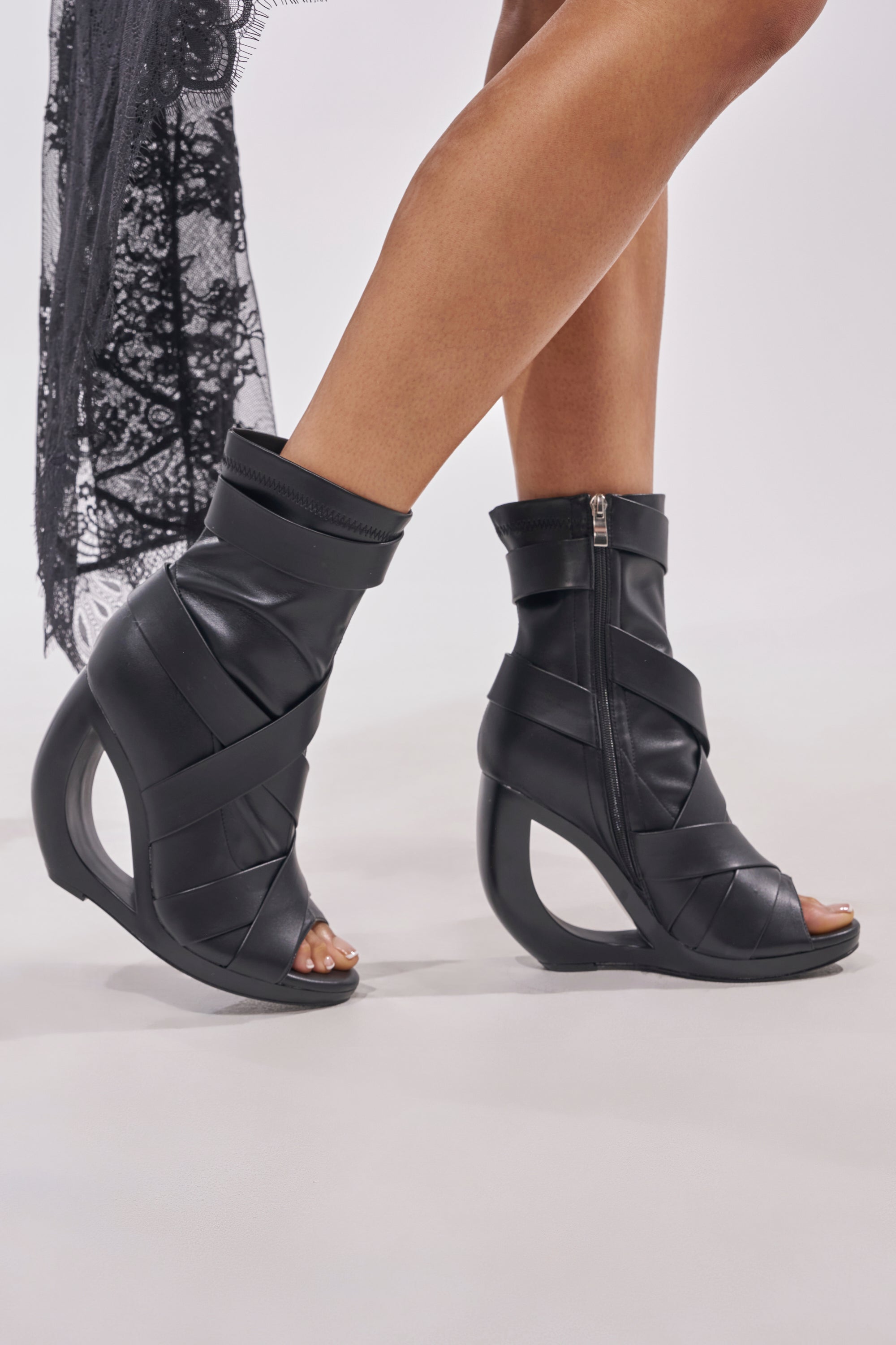 A person wears the AZALEA WANG IGNITE Black Wedge Open Toe Bootie with belting detail, featuring a futuristic design, geometric cutout heel, and crisscross straps, walking on a light background with black lace visible. 1