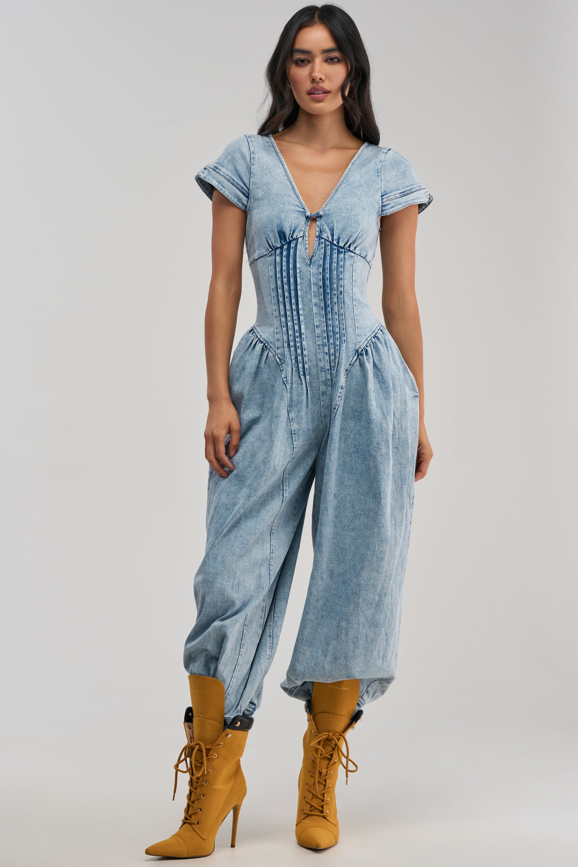 Wearing the MUST HAVE SHORT SLEEVE DENIM JUMPSUIT, which features a corset-style bodice and voluminous pant legs in a light wash, she pairs it with mustard yellow lace-up heeled boots while posing against a plain background for a chic streetwear-inspired look.