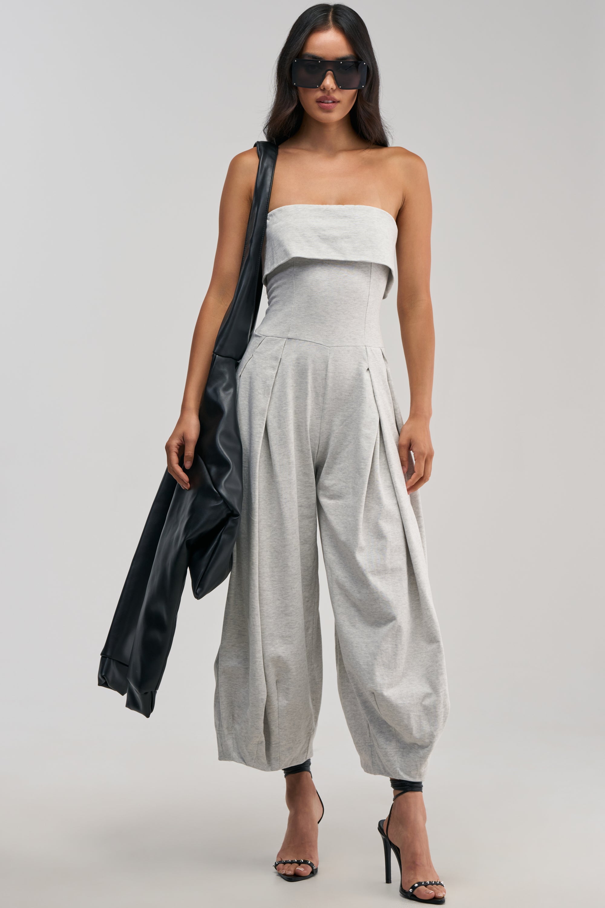 A woman stands against a plain light background wearing the COMFY AND CUTE STRAPLESS JUMPSUIT IN HEATHER GREY, which features wide, pleated legs. She pairs the jumpsuit with black high heels and dark sunglasses, and carries a black jacket over her shoulder.