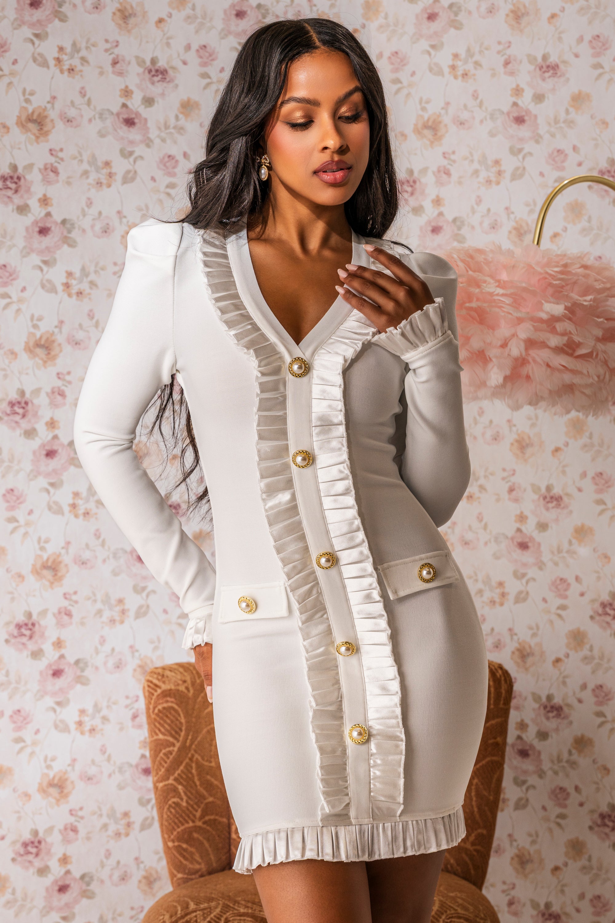 A woman poses elegantly against floral wallpaper in the SAKURA BANDAGE RUFFLE MINI DRESS IN WHITE, a fitted mini dress featuring gold buttons and satin ruffle trim—perfect for bridal events. 3