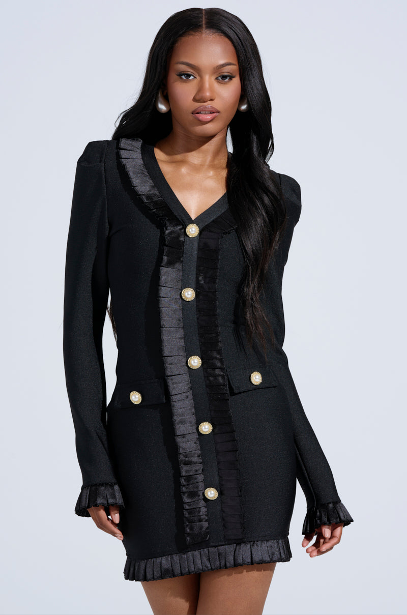 A woman wearing the SAKURA BANDAGE RUFFLE MINI DRESS with long sleeves, pearl-embellished buttons, and pleated satin trim stands against a plain light background, looking confidently at the camera.