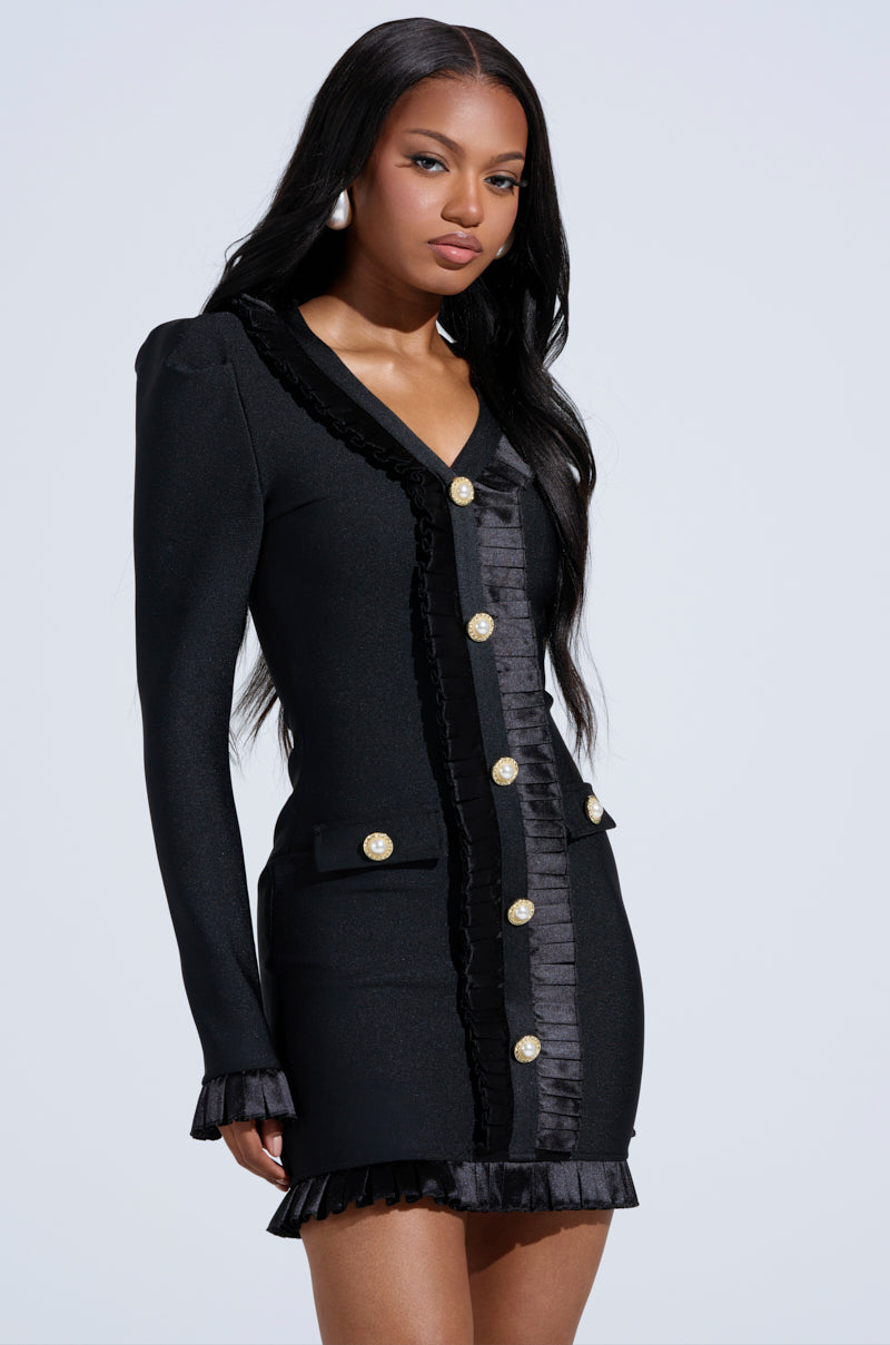 A woman with long black hair models the SAKURA BANDAGE RUFFLE MINI DRESS, a fitted black long-sleeve dress with pearl-embellished buttons and ruffled trim, standing against a plain light background.