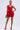 A woman stands confidently in the FELT CUTE RUFFLE BANDAGE MINI DRESS—a short, sleeveless bandage dress featuring a sculpted silhouette and ruffled layers at the hips. She accessorizes with a clutch, red strappy heels, colorful earrings, and has her hair pulled back.