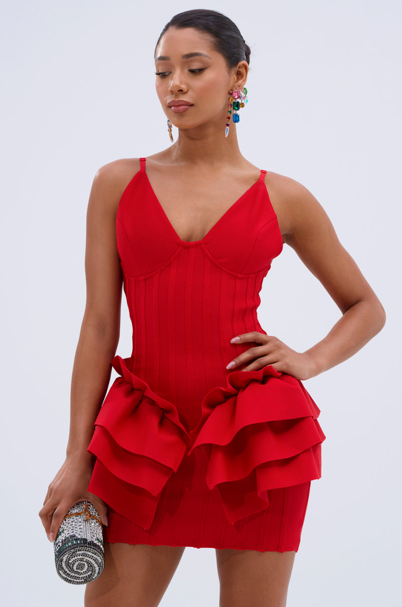 A woman stands against a light background wearing the FELT CUTE RUFFLE BANDAGE MINI DRESS, a fitted red mini dress with ruffled layers at the hips. She holds a beaded clutch, wears statement earrings, and has her hair tied back.